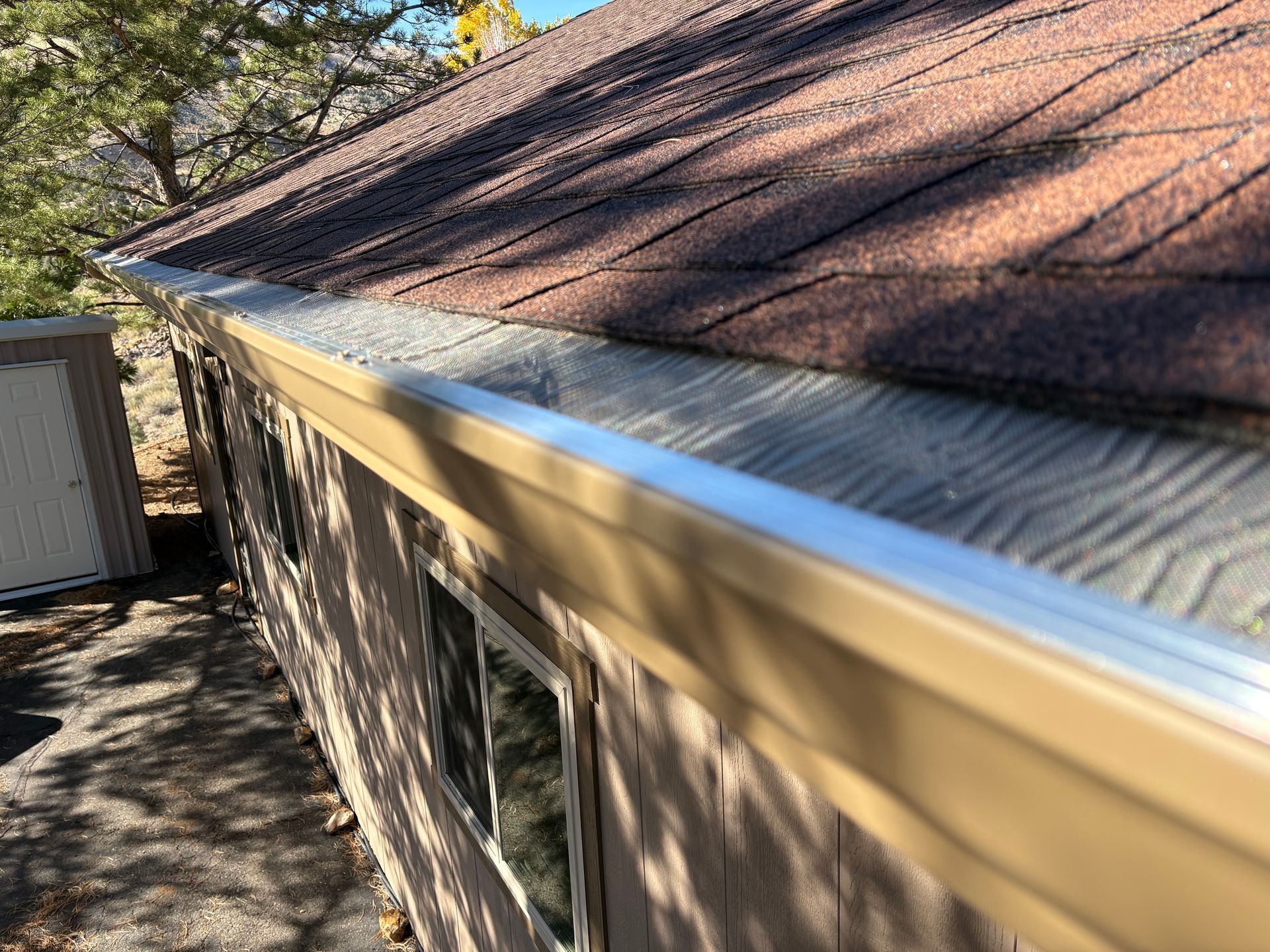 Beige gutter with a mesh guard on a brown shingle roof, attached to a light brown building.
