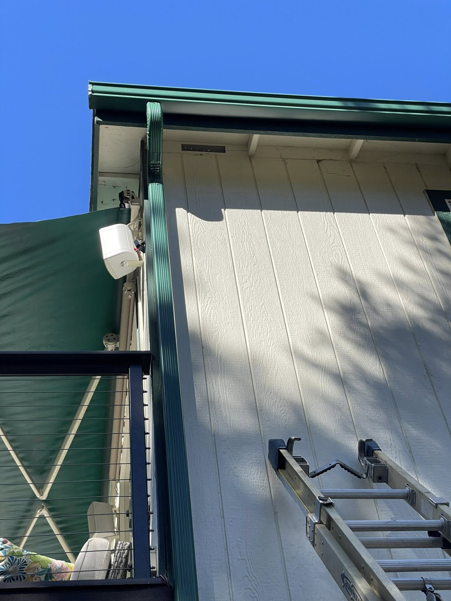 White building with green trim, a ladder, and a white device attached under the roof. Sunny day.