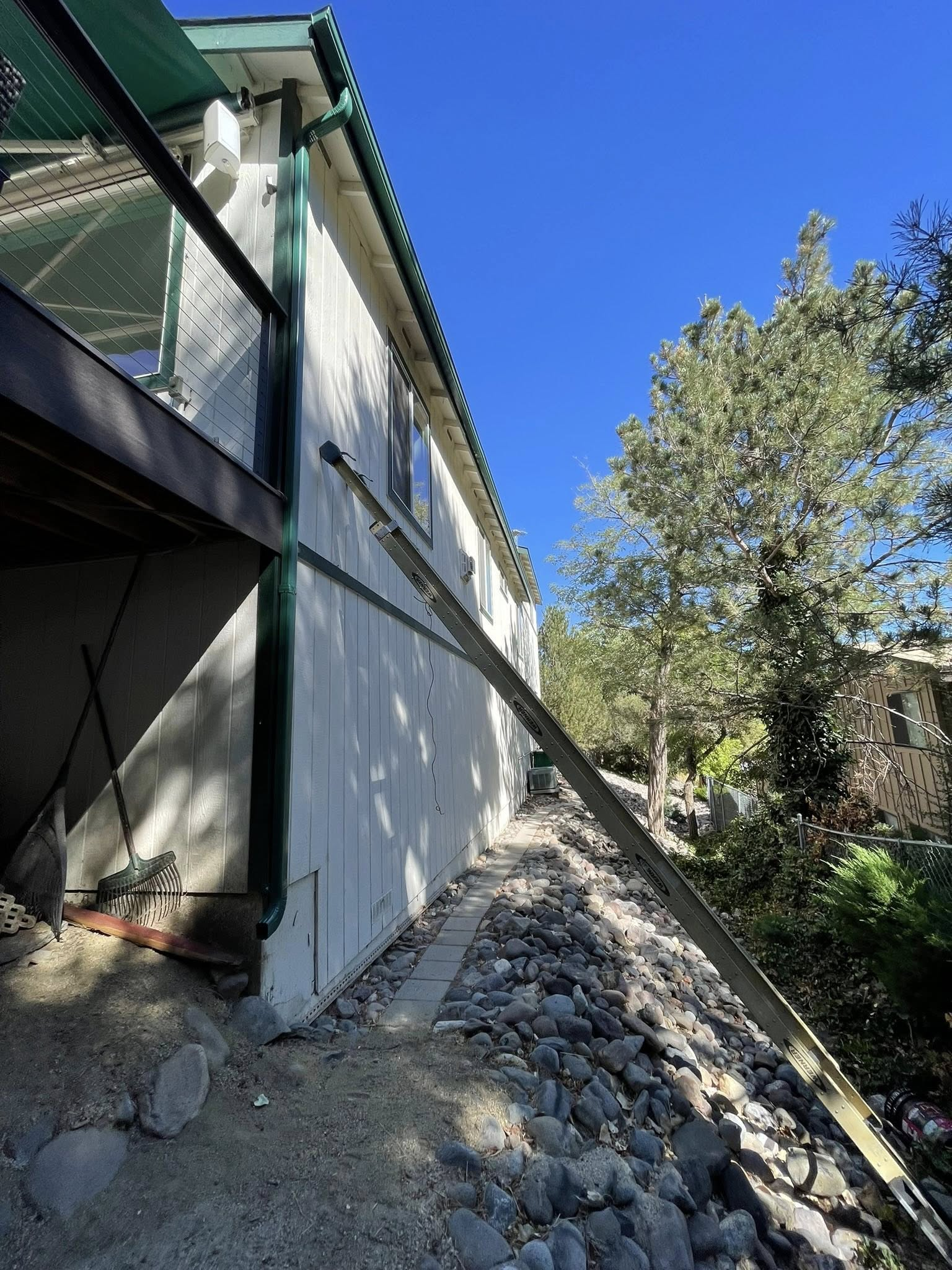 Side of a white house with a green trim, a retaining wall, and a ladder leaning against the siding on a sunny day.