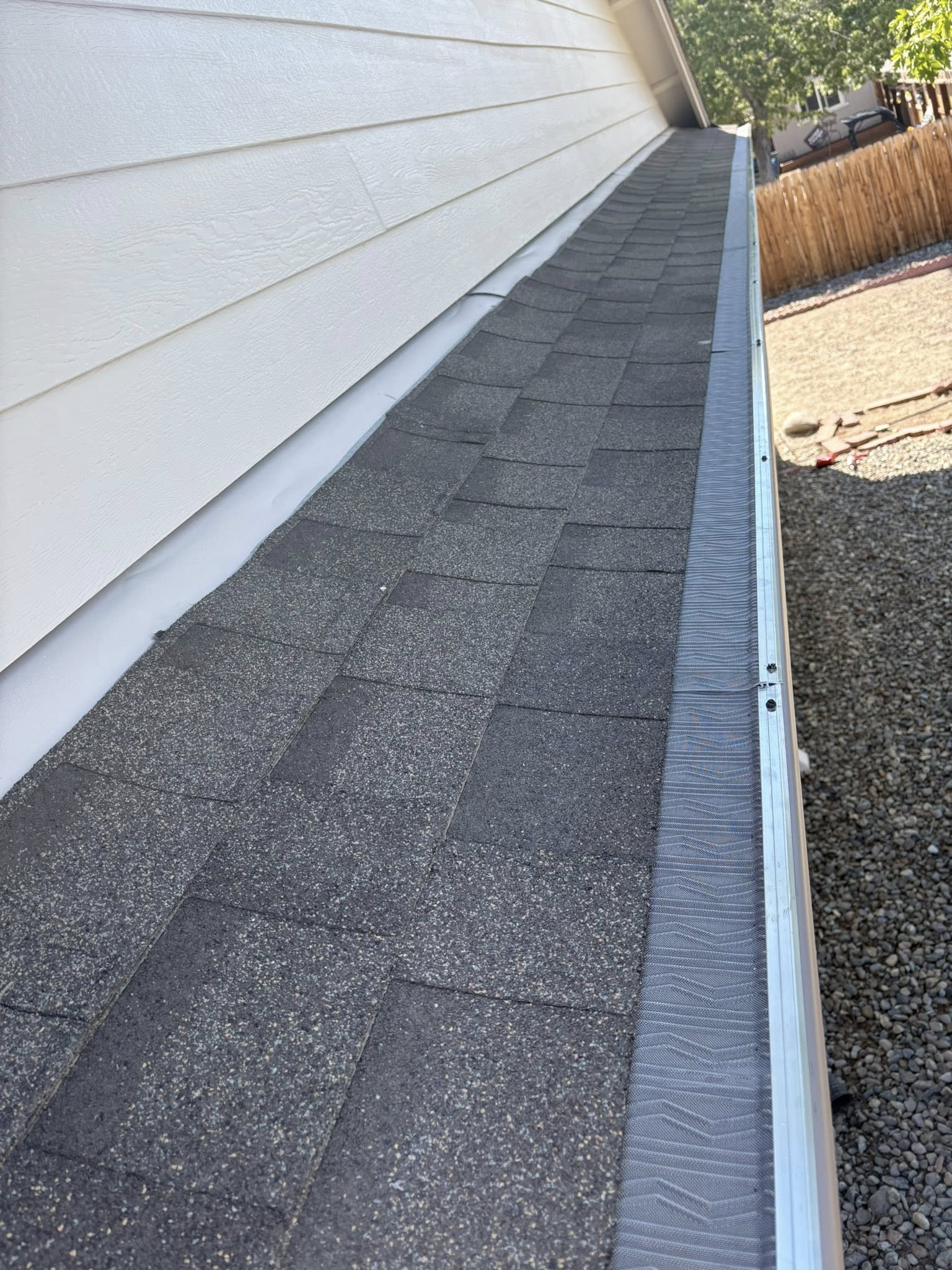 A close-up of a roof edge with asphalt shingles, white siding, and a gutter, on a sunny day.