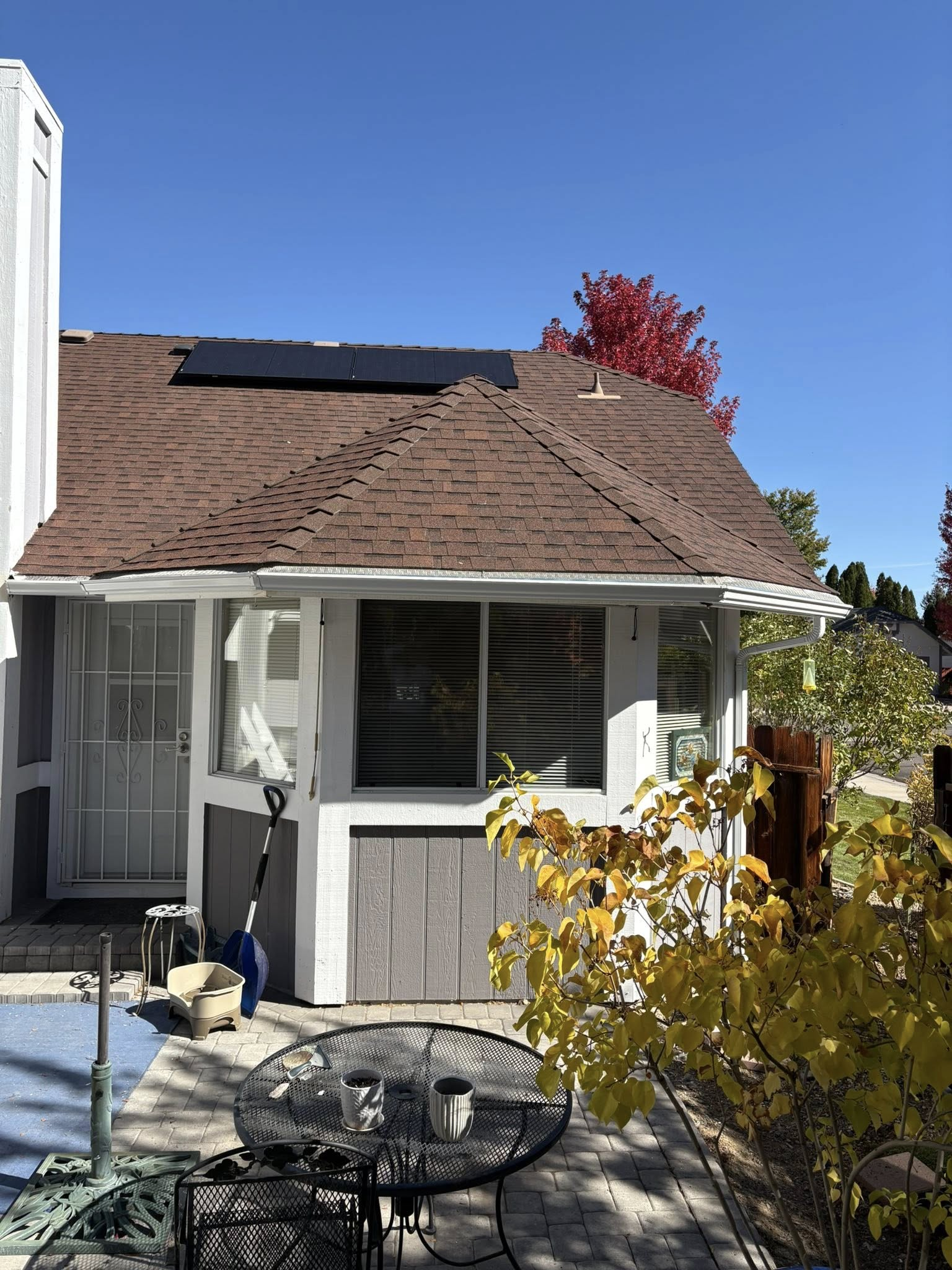 House exterior with solar panels on roof, dark panels, patio furniture, and colorful autumn foliage under blue sky.