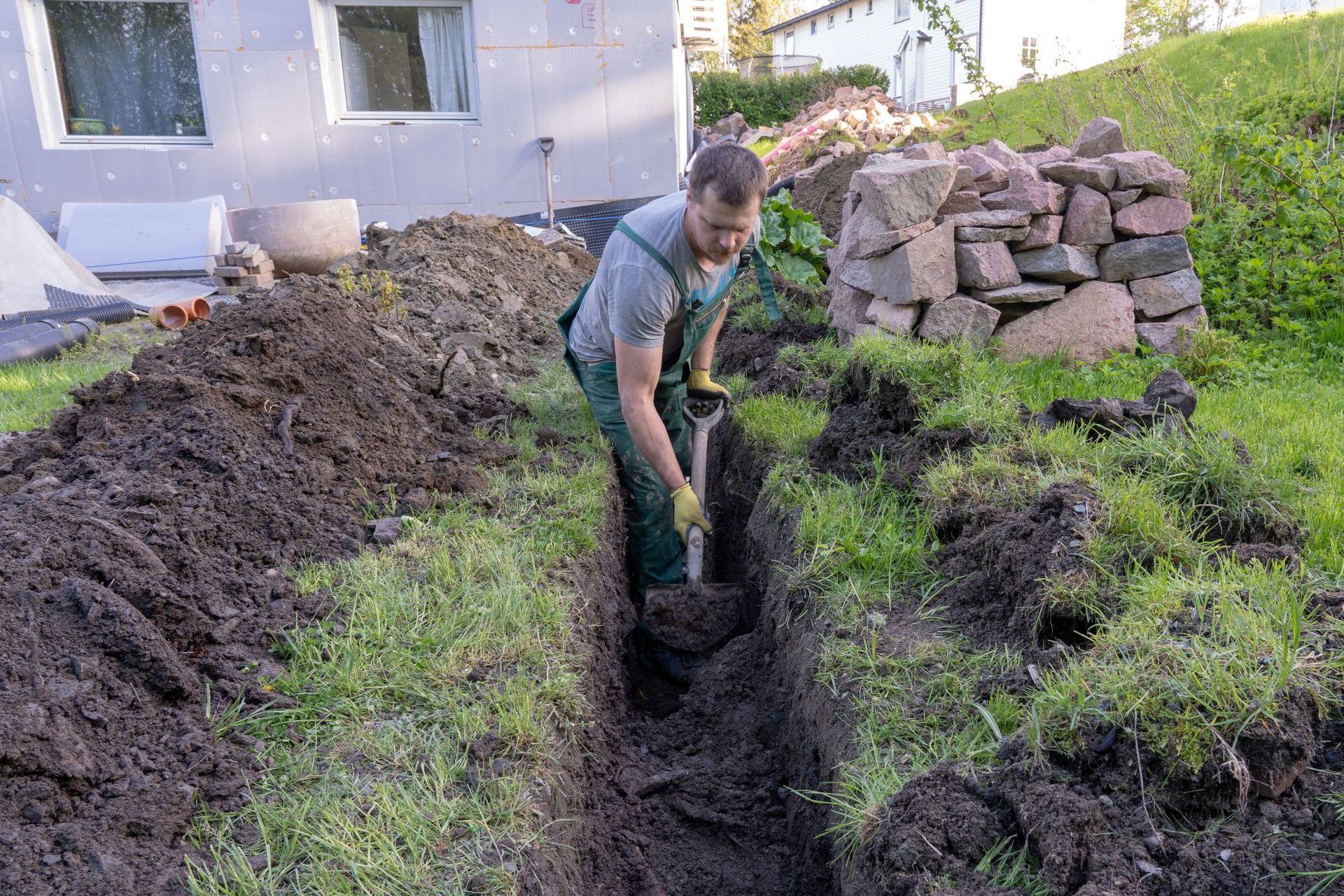 Man digging a trench in grassy yard near a building, using a shovel.