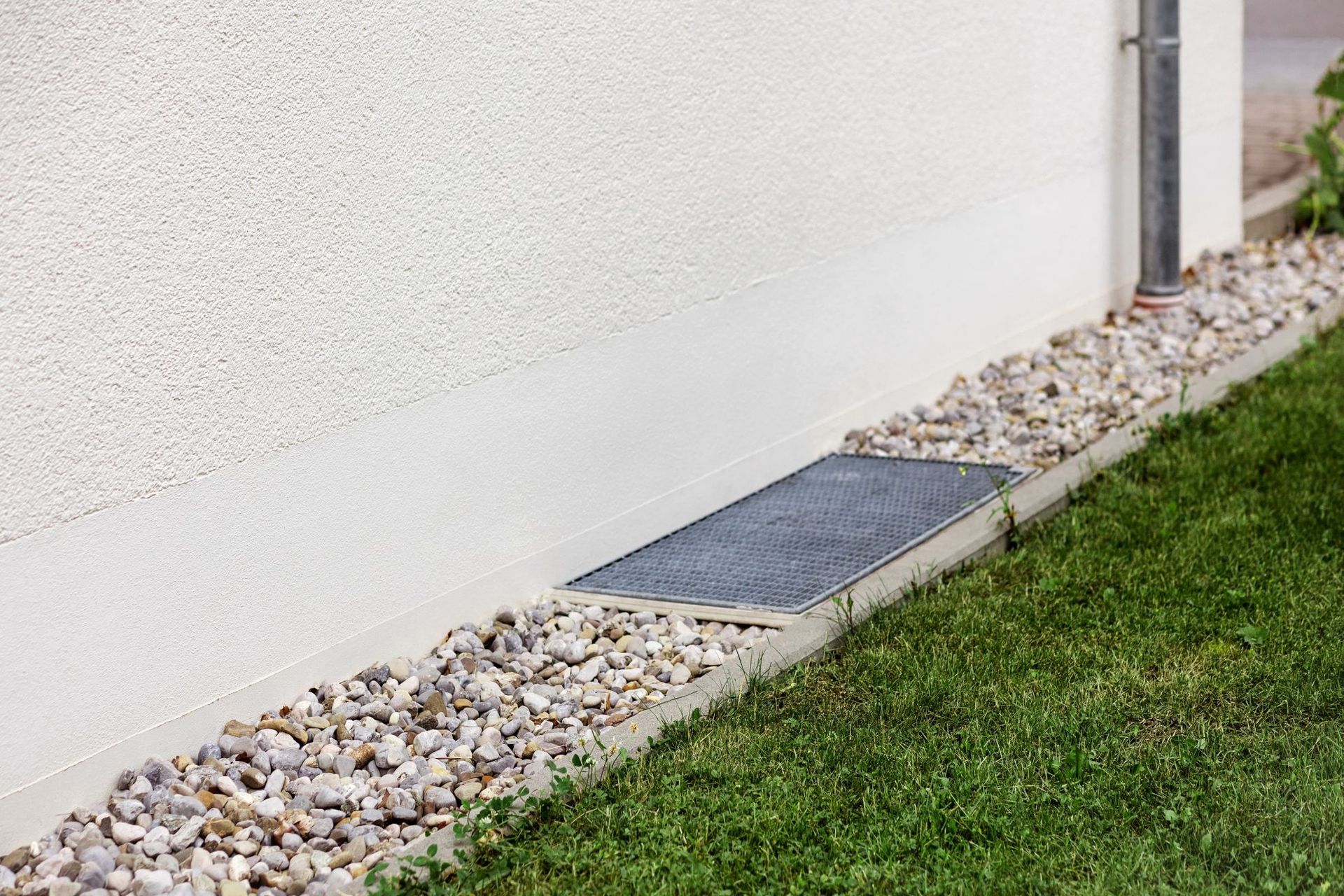 Concrete foundation with gravel and a grate at the base of a white stucco wall next to a grassy lawn.