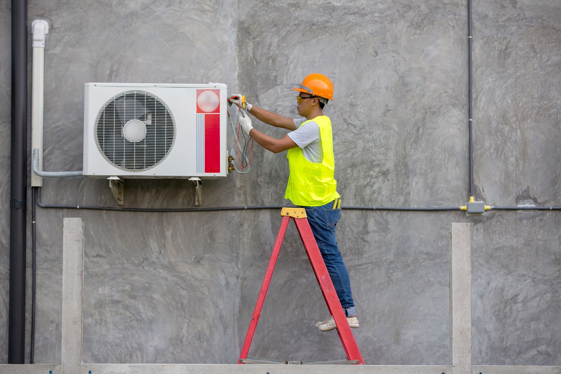 A man is standing on a ladder fixing an air conditioner on the side of a building.