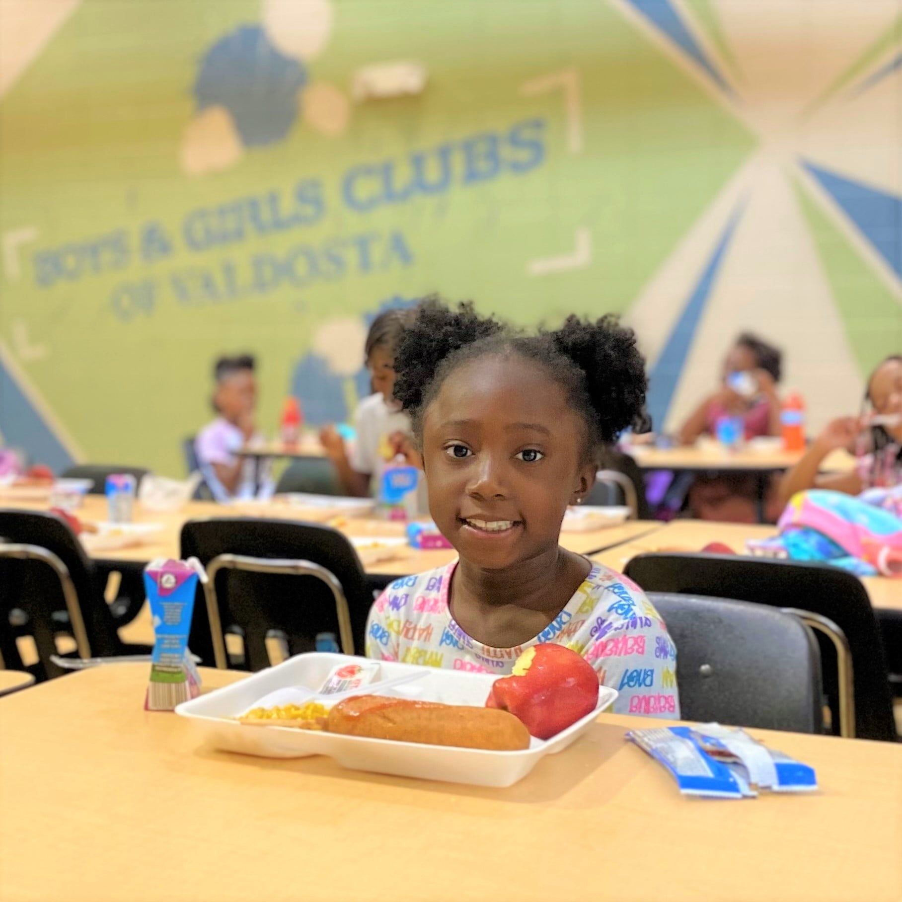 A young girl smiles at the camera, seated at a table in a Boys & Girls Club. She has a lunch tray with food.