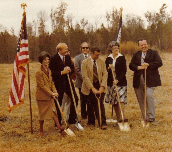 People with shovels at a groundbreaking ceremony, outdoors, holding shovels in a brown dirt field. Flags in the background.