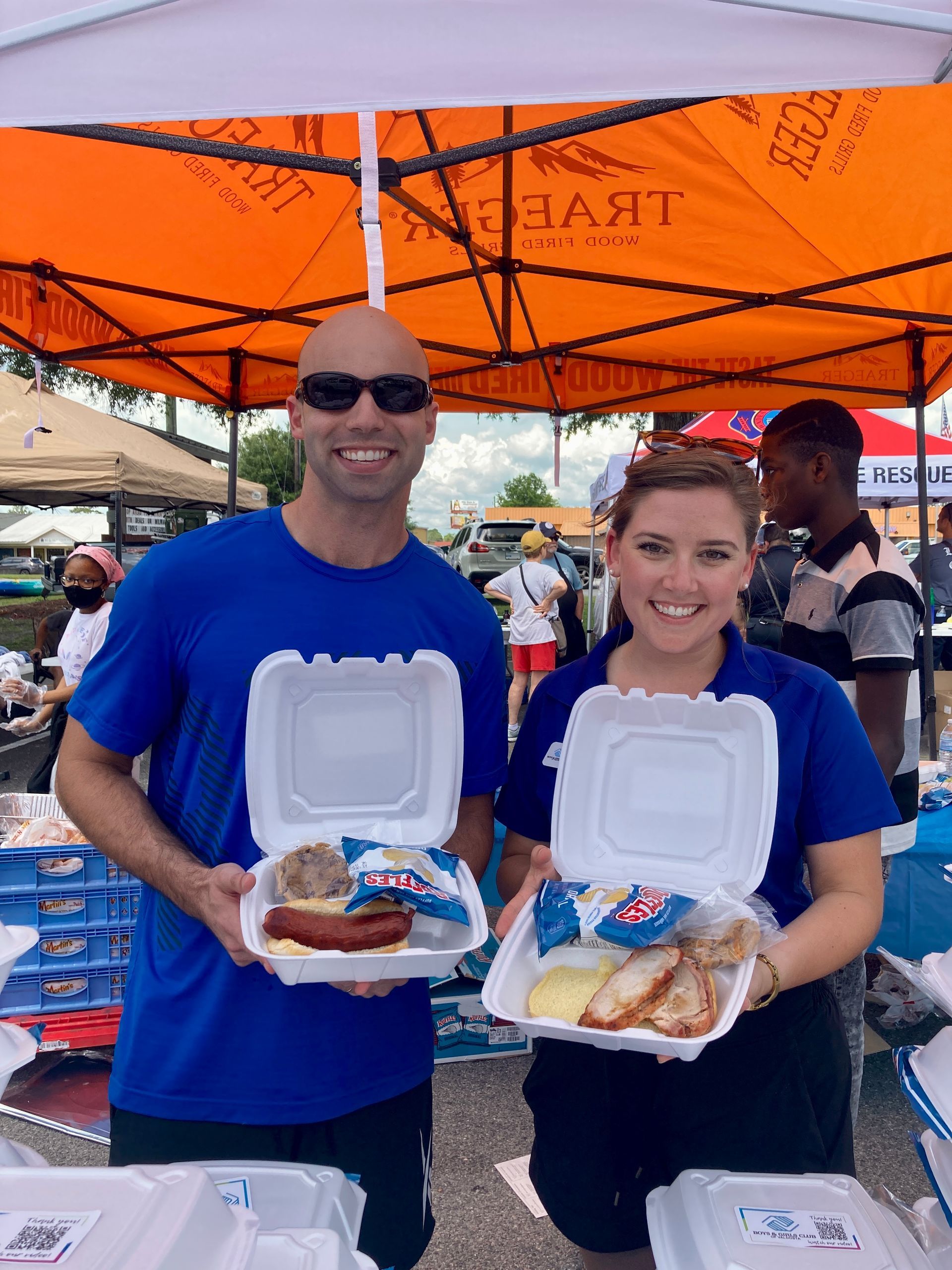 Two people smiling, holding meals in styrofoam containers under an orange tent.