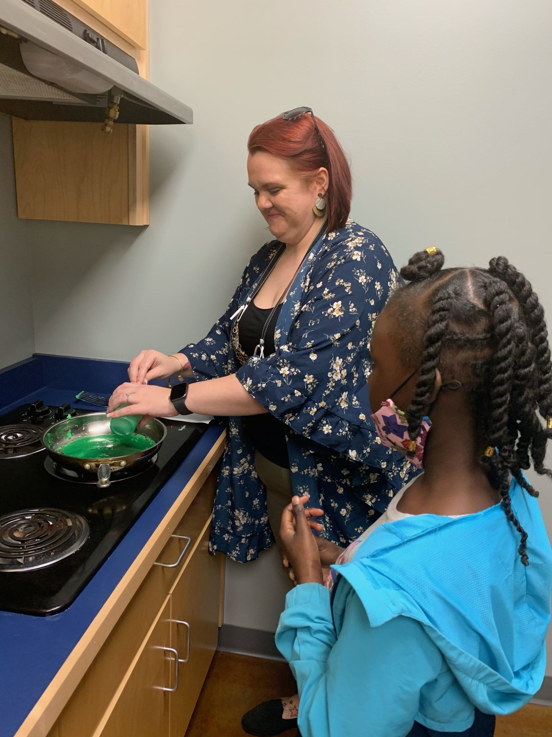 Woman and girl cooking together on stovetop; green food in pan.