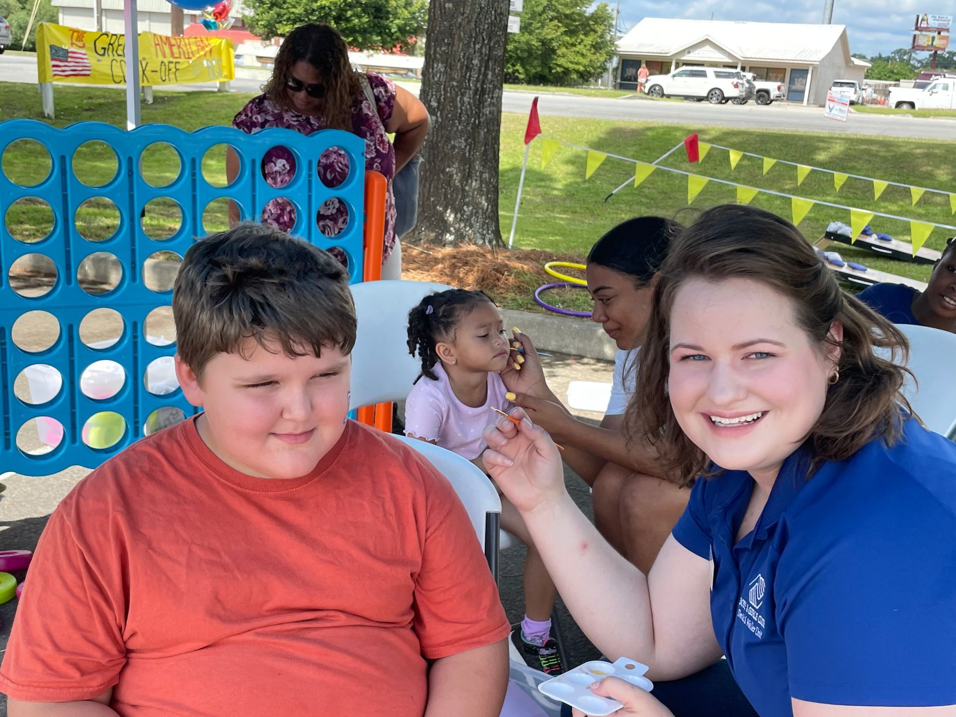 Boy smiles as another person applies face paint at outdoor event; Connect Four game in the background.