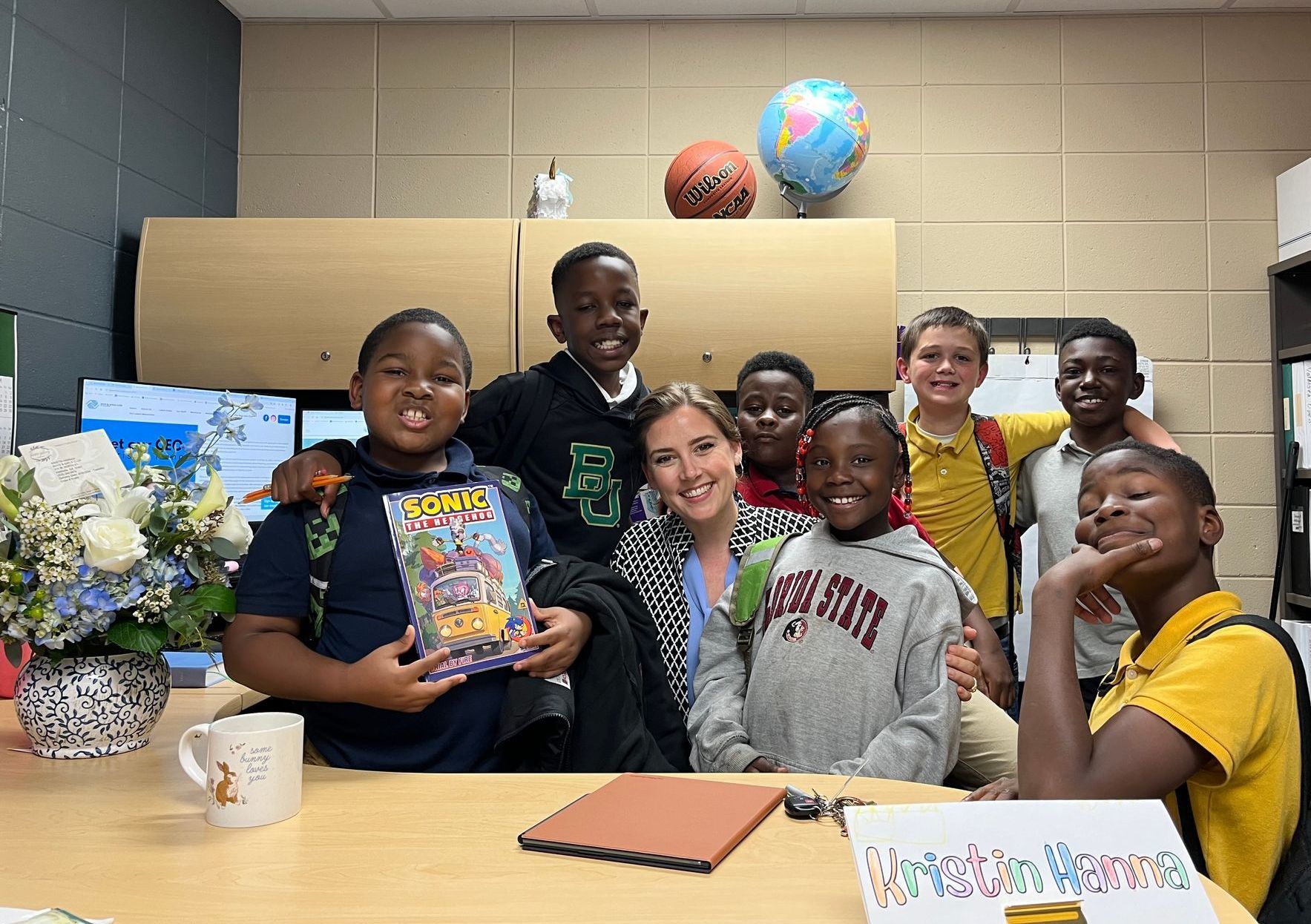 CEO smiles with students. They're at a desk with a globe, basketball, and flowers in an office.