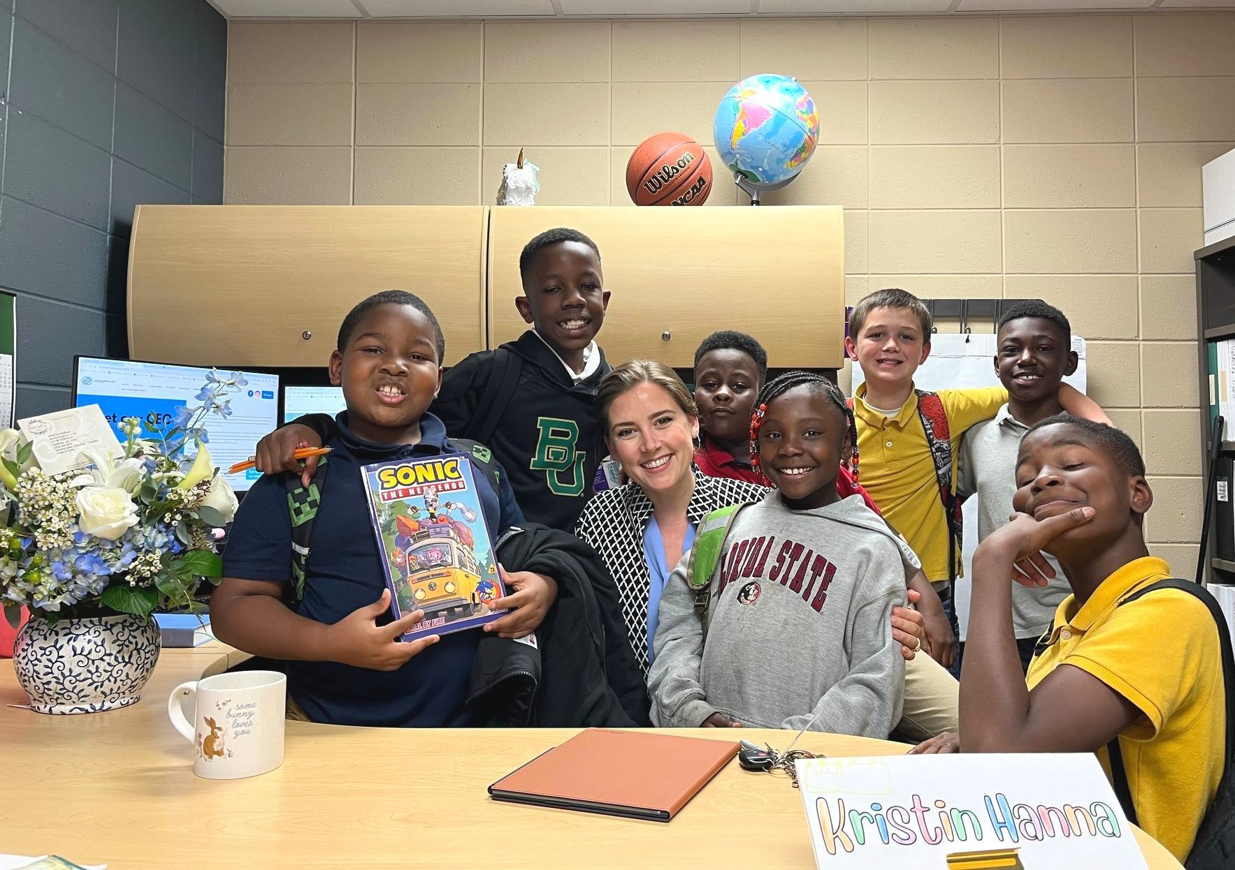 CEO smiles with students. They're at a desk with a globe, basketball, and flowers in an office.