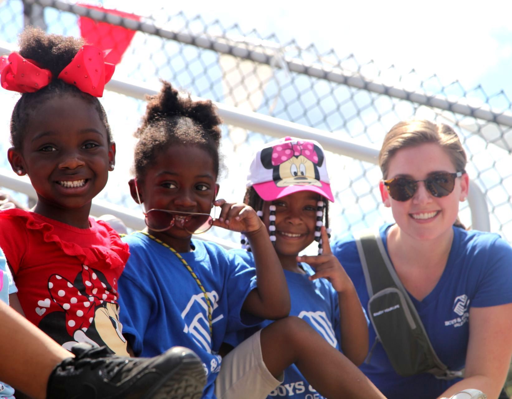 Four smiling children and an adult pose outdoors; a girl in a bow smiles next to others in blue shirts.