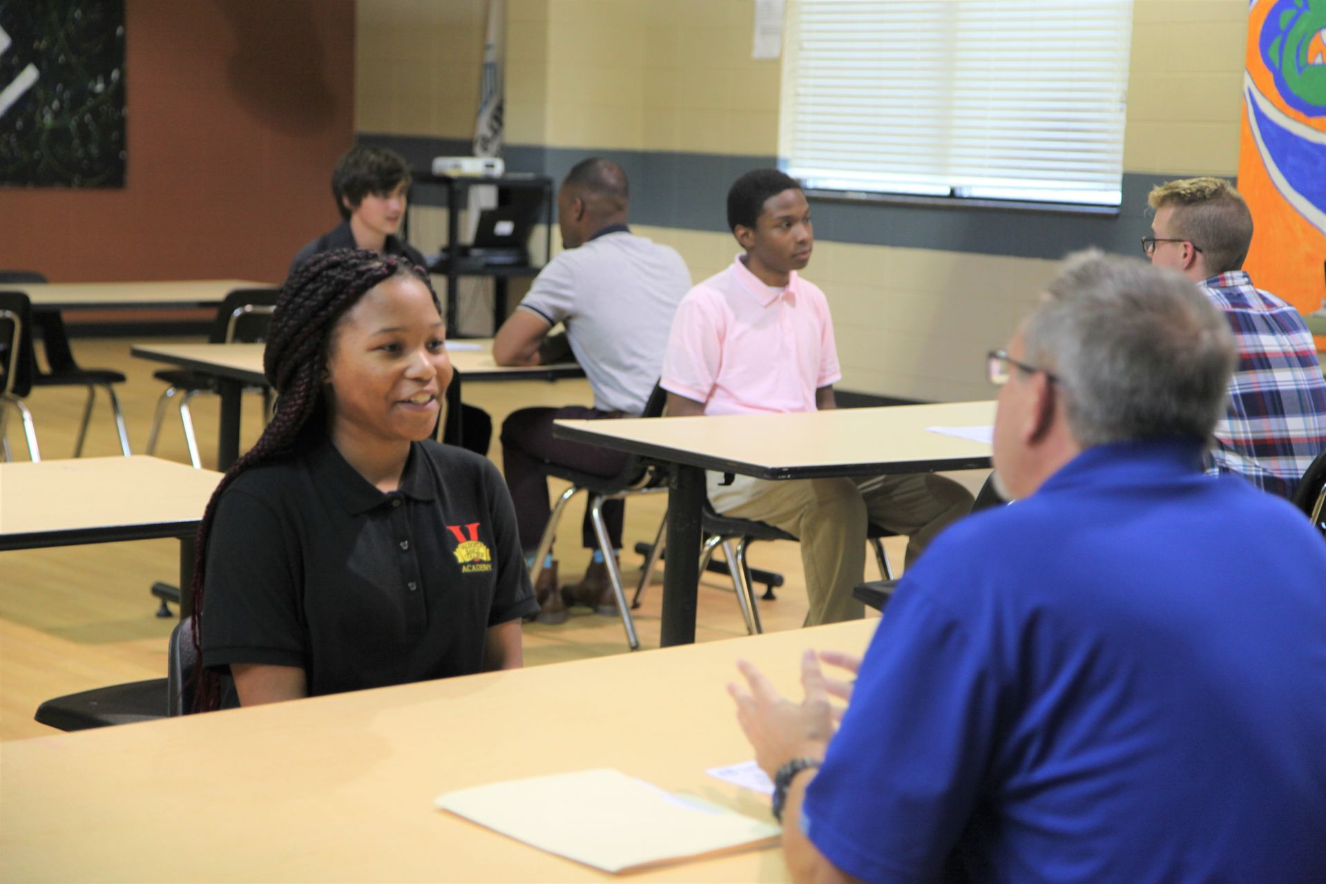 A student in a black shirt is being interviewed at a table; others wait at desks.