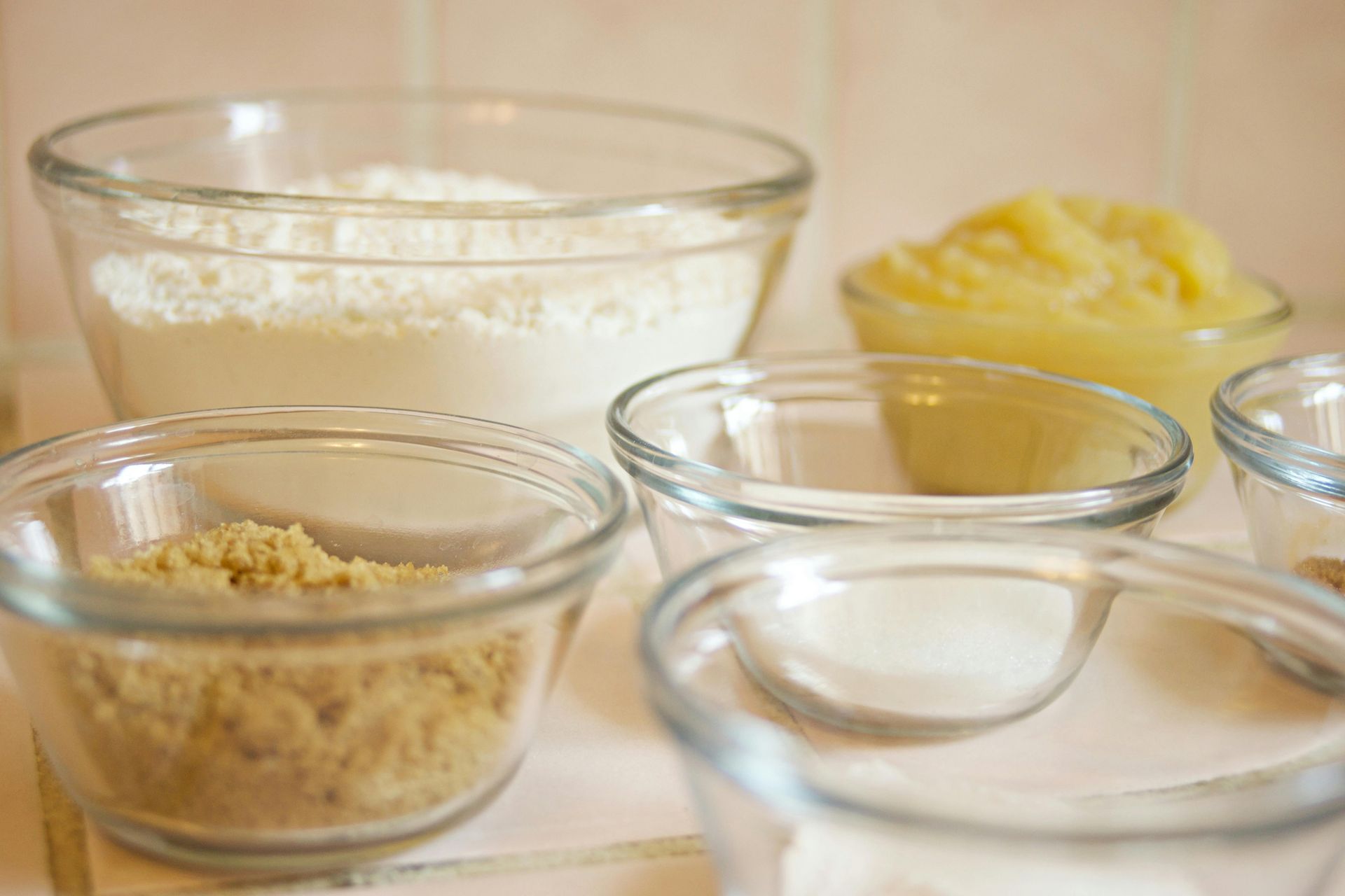 Glass bowls filled with baking ingredients, including flour, brown sugar, and apple sauce, arranged on a kitchen counter.