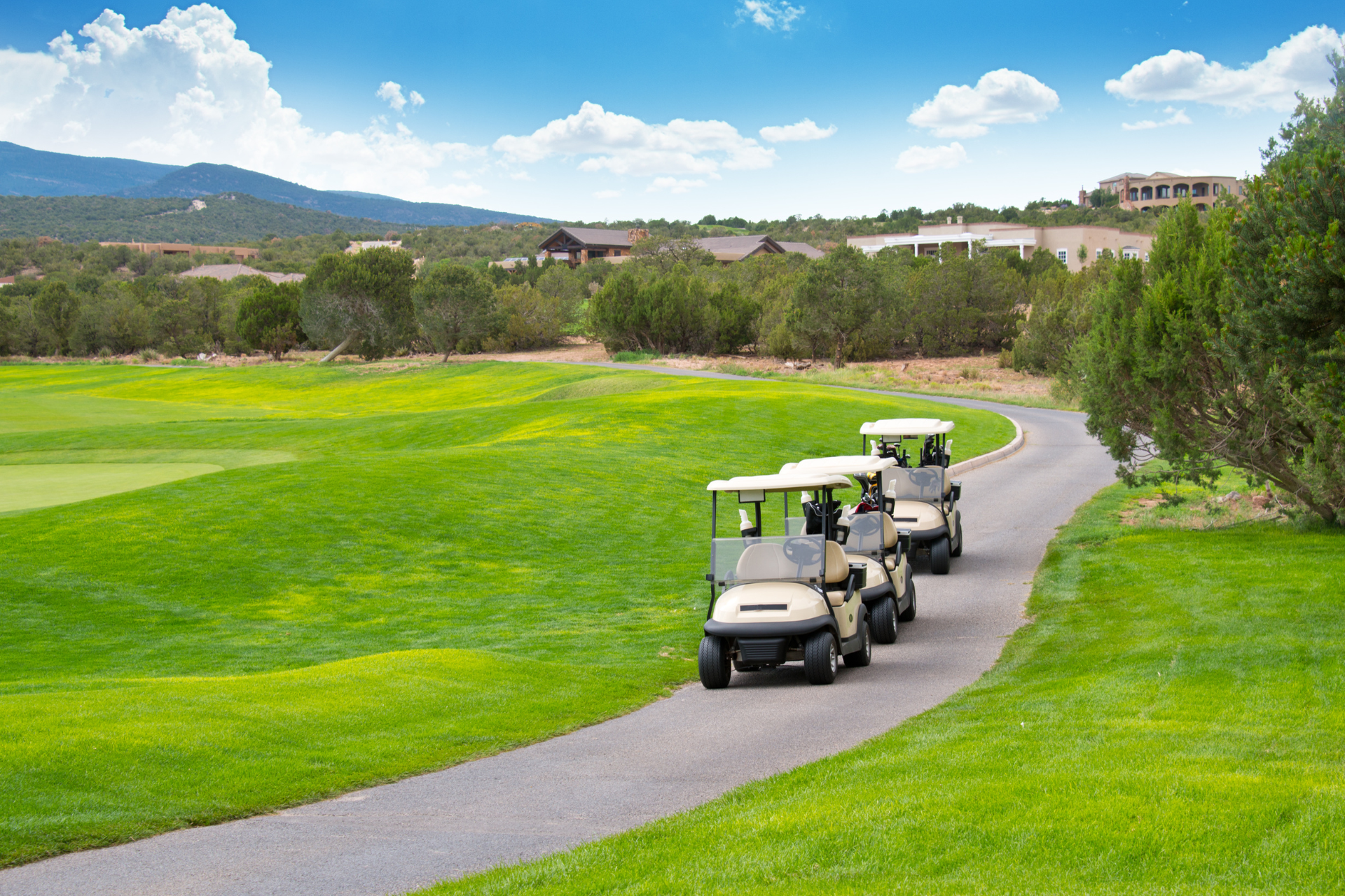 Three golf carts on a paved path through a green golf course, mountains in the background.