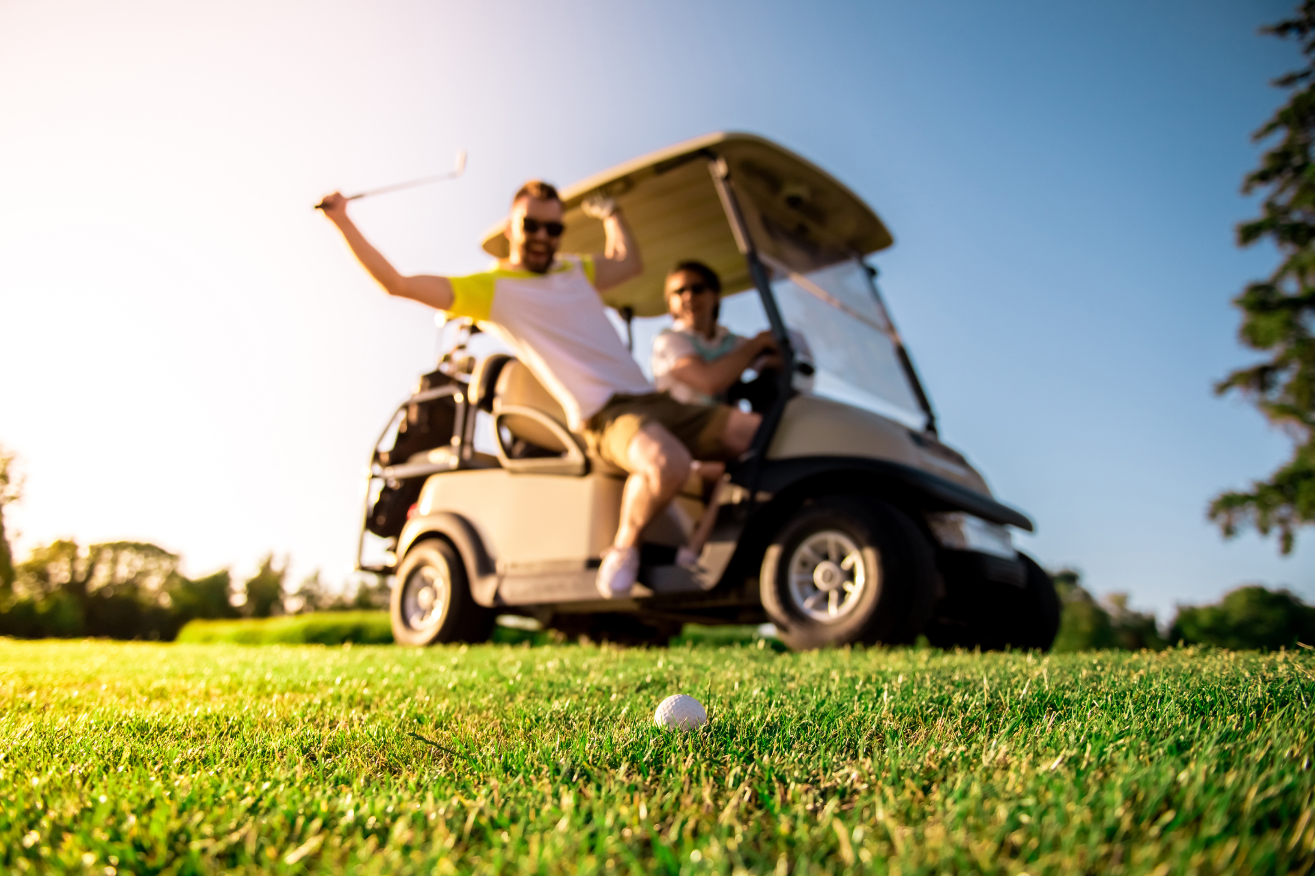 Two men in a golf cart on a green, one swings a club. Sunny, blue sky.