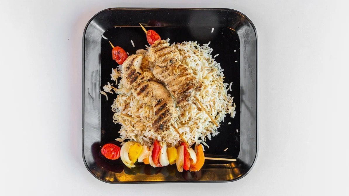 Grilled chicken and rice on a black plate, garnished with a skewer of vegetables and chili peppers.
