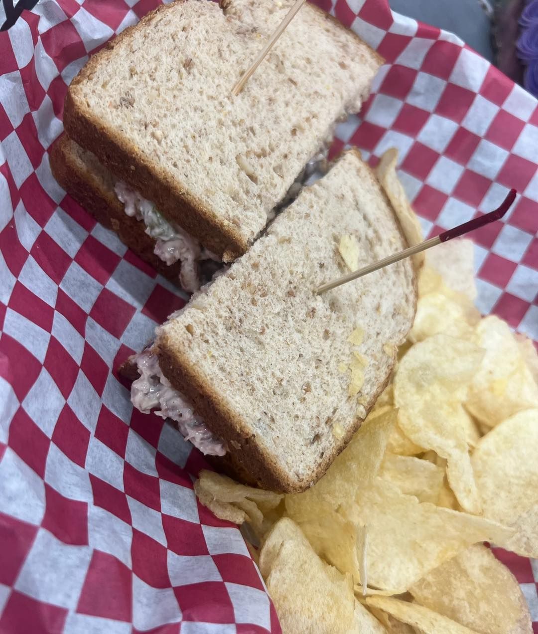 Sandwich on wheat bread, cut in half and held together with a toothpick, with potato chips in a checkered basket.