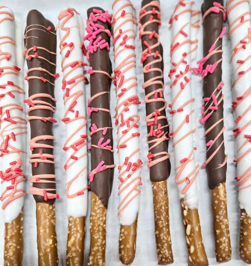 Chocolate-covered pretzel rods, drizzled with pink and decorated with sprinkles, arranged in a row.