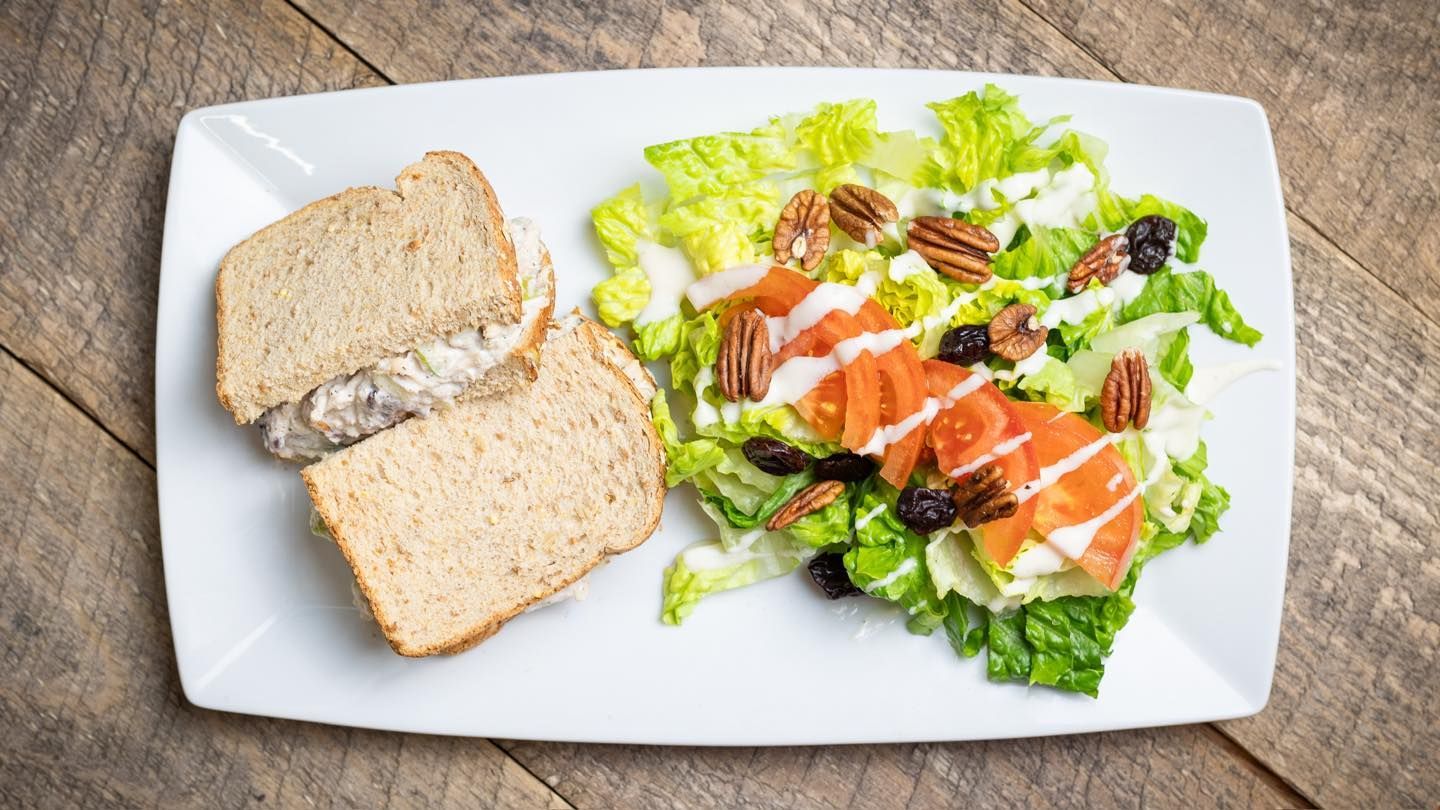 Sandwich and salad on a white plate atop a wooden surface. Salad has pecans and tomatoes.