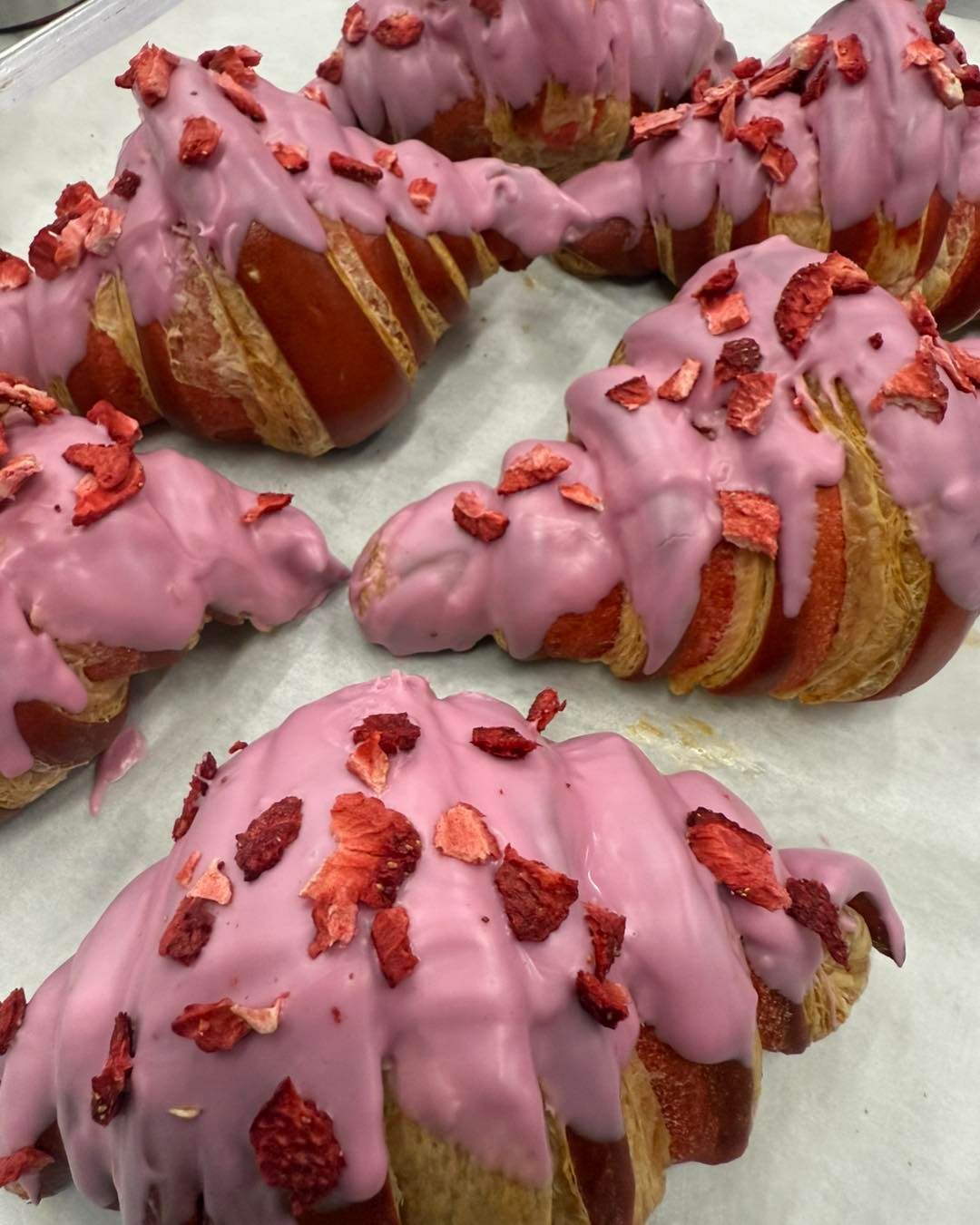 Strawberry-glazed croissants, topped with dried strawberry pieces, on a light-colored surface.