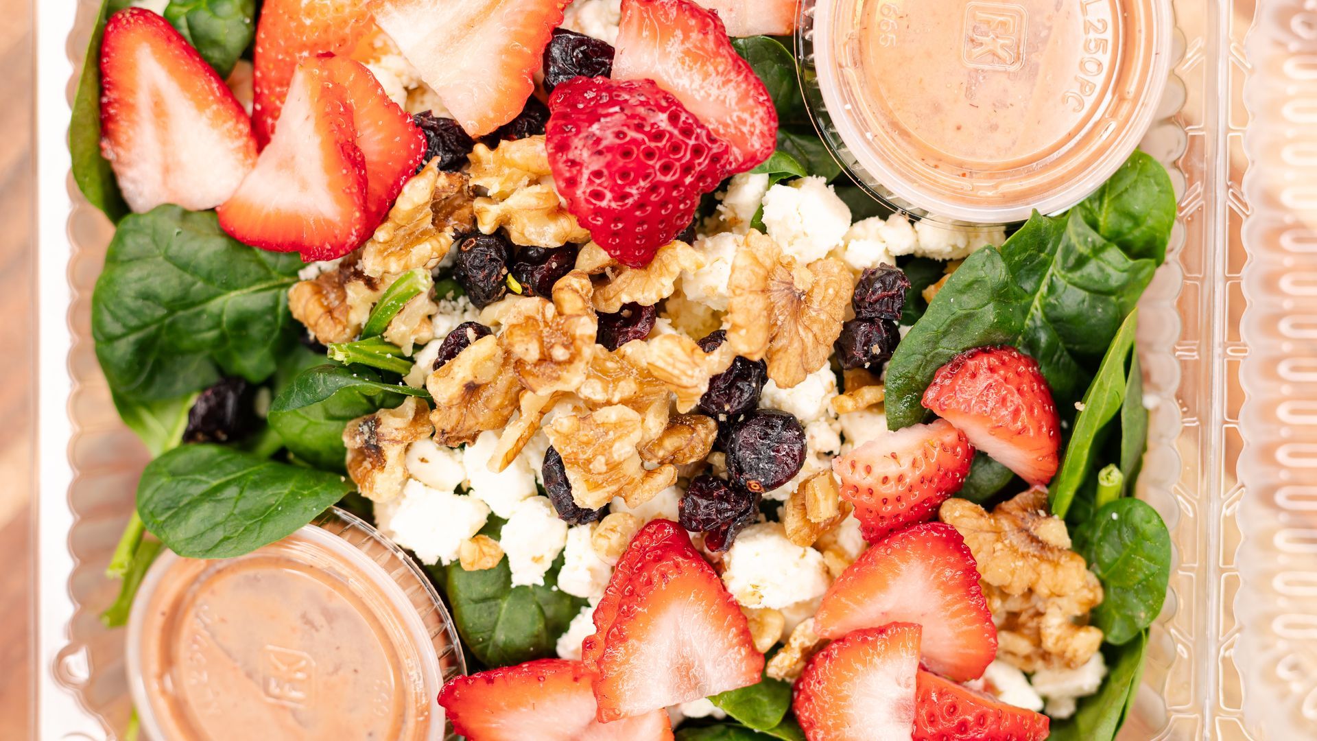 Salad in clear container: spinach, strawberries, walnuts, raisins, feta cheese, with two containers of dressing.