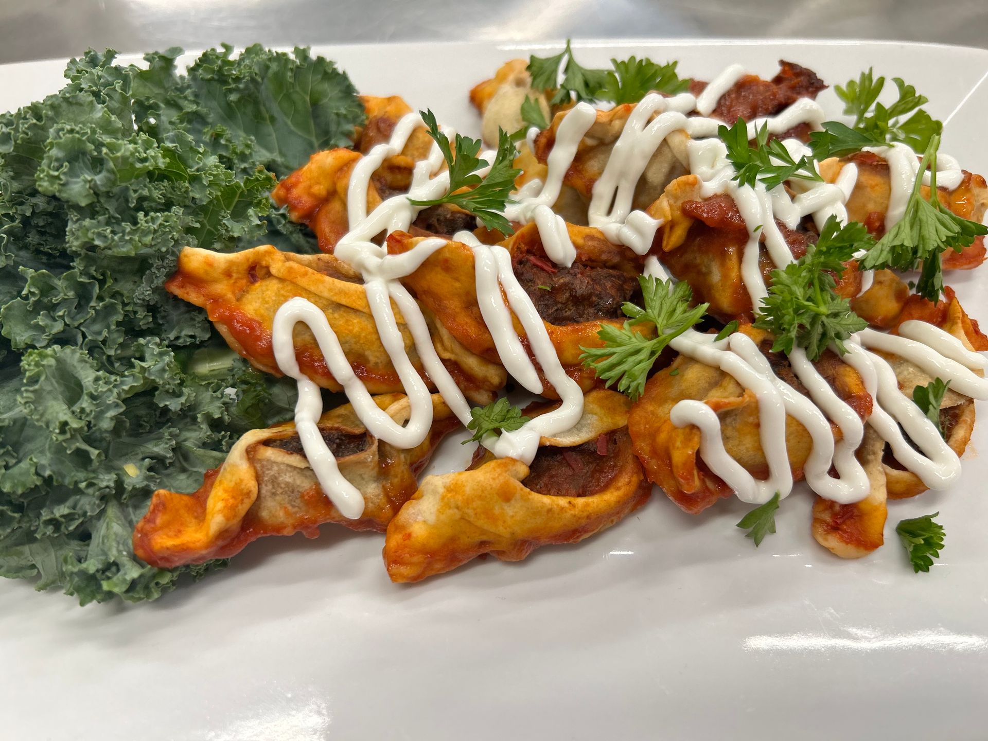 Fried pastry with a red sauce, drizzled with white cream, garnished with parsley, and kale on a white plate.