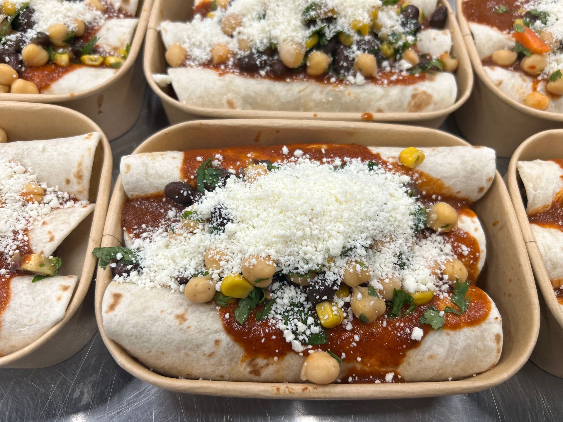 Enchiladas in rectangular containers, topped with sauce, chickpeas, corn, cilantro, and shredded cheese.