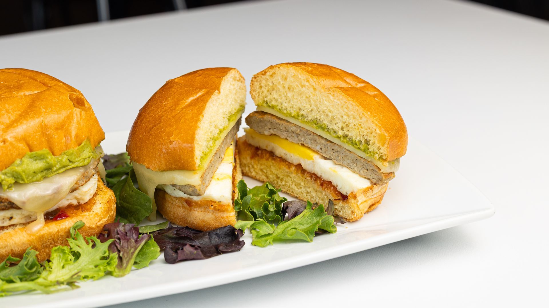 Three breakfast sliders with fried egg, cheese, and pesto on a white plate.