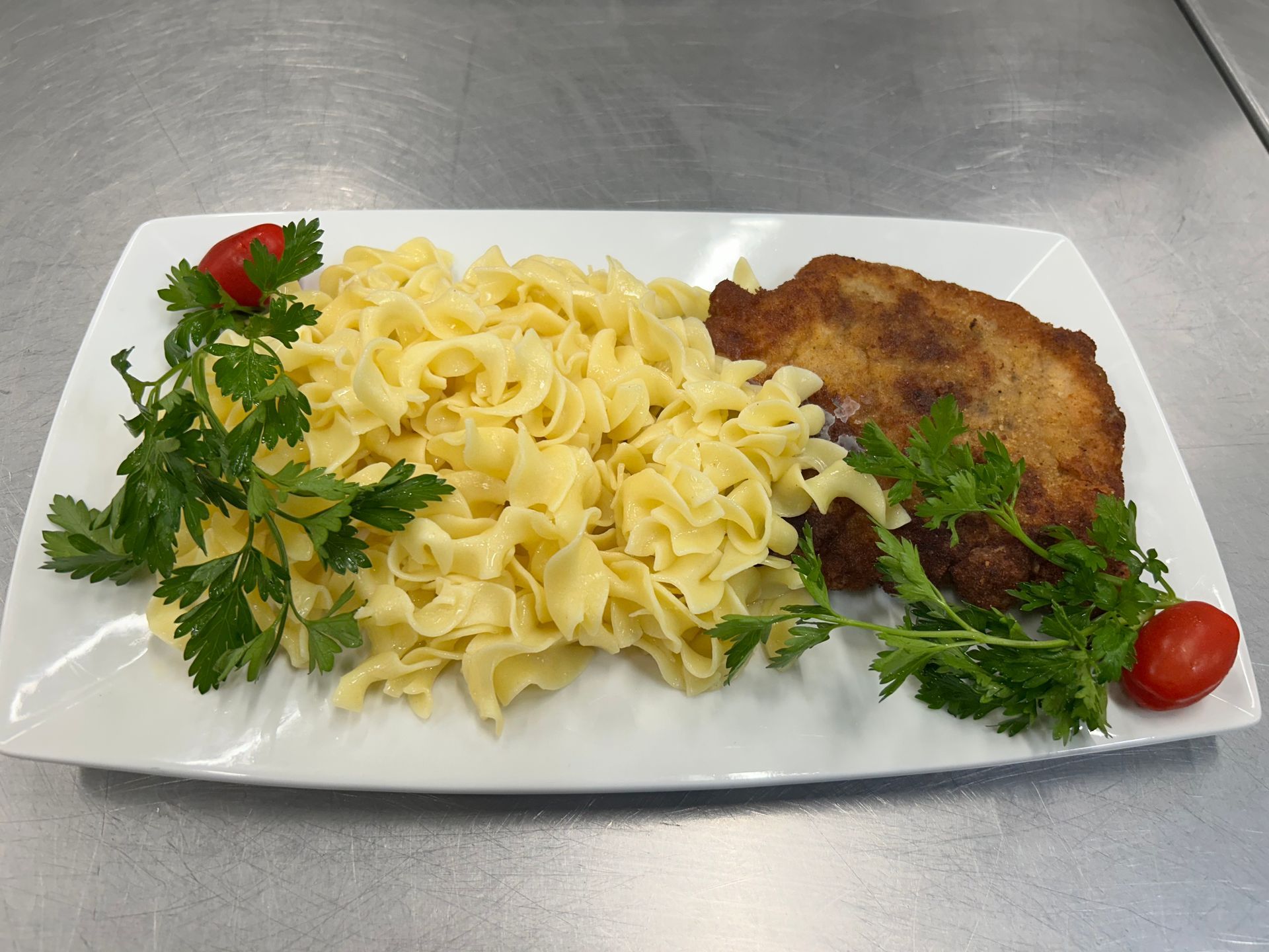 Fried breaded cutlet with noodles, parsley garnish, and tomatoes on a white rectangular plate.