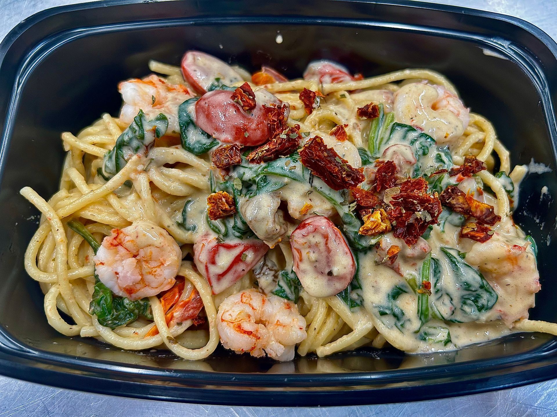 Pasta dish with shrimp, spinach, tomatoes, and creamy sauce in a black container.