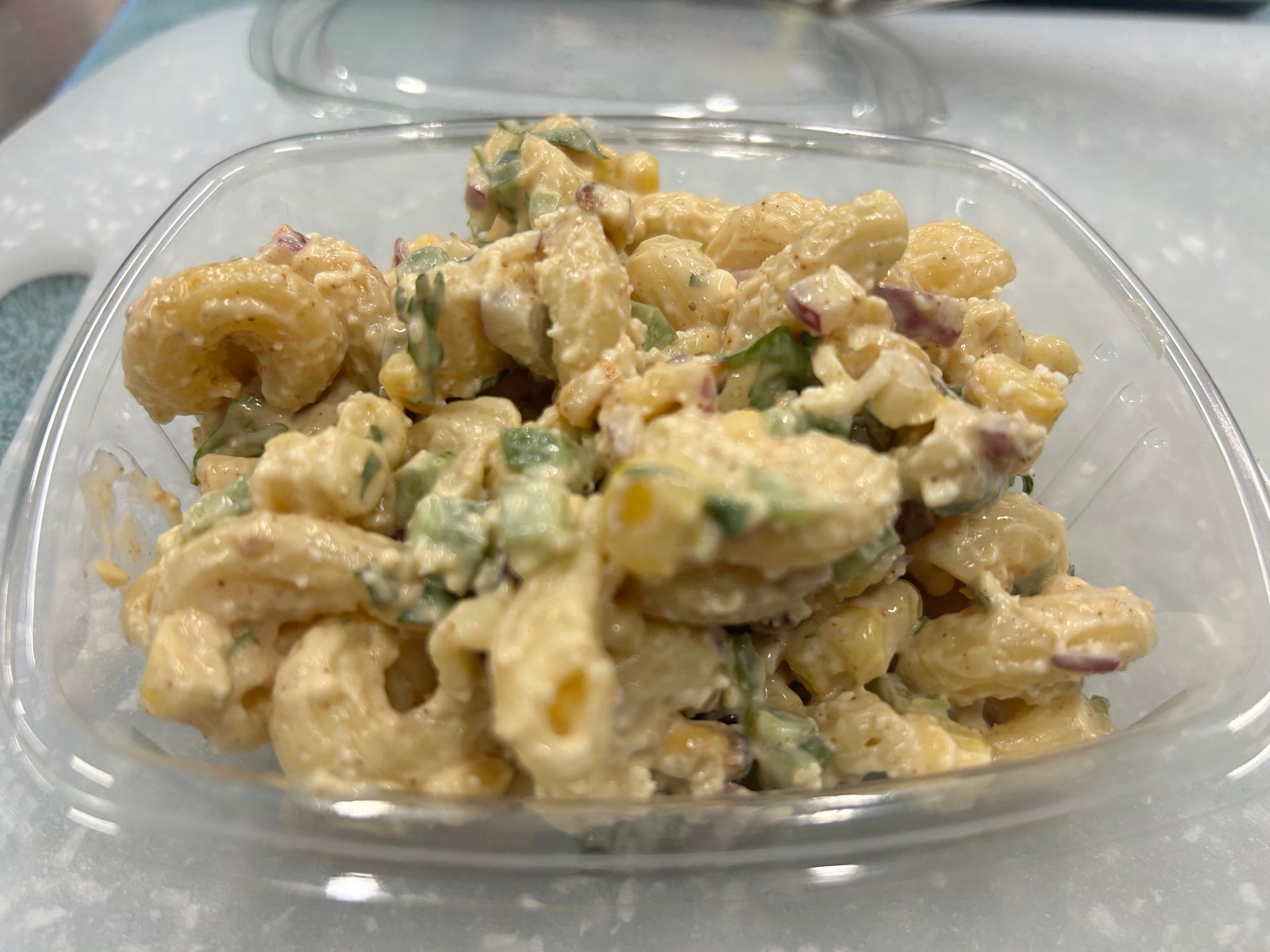Macaroni salad in a clear plastic container; includes macaroni, vegetables, and dressing.