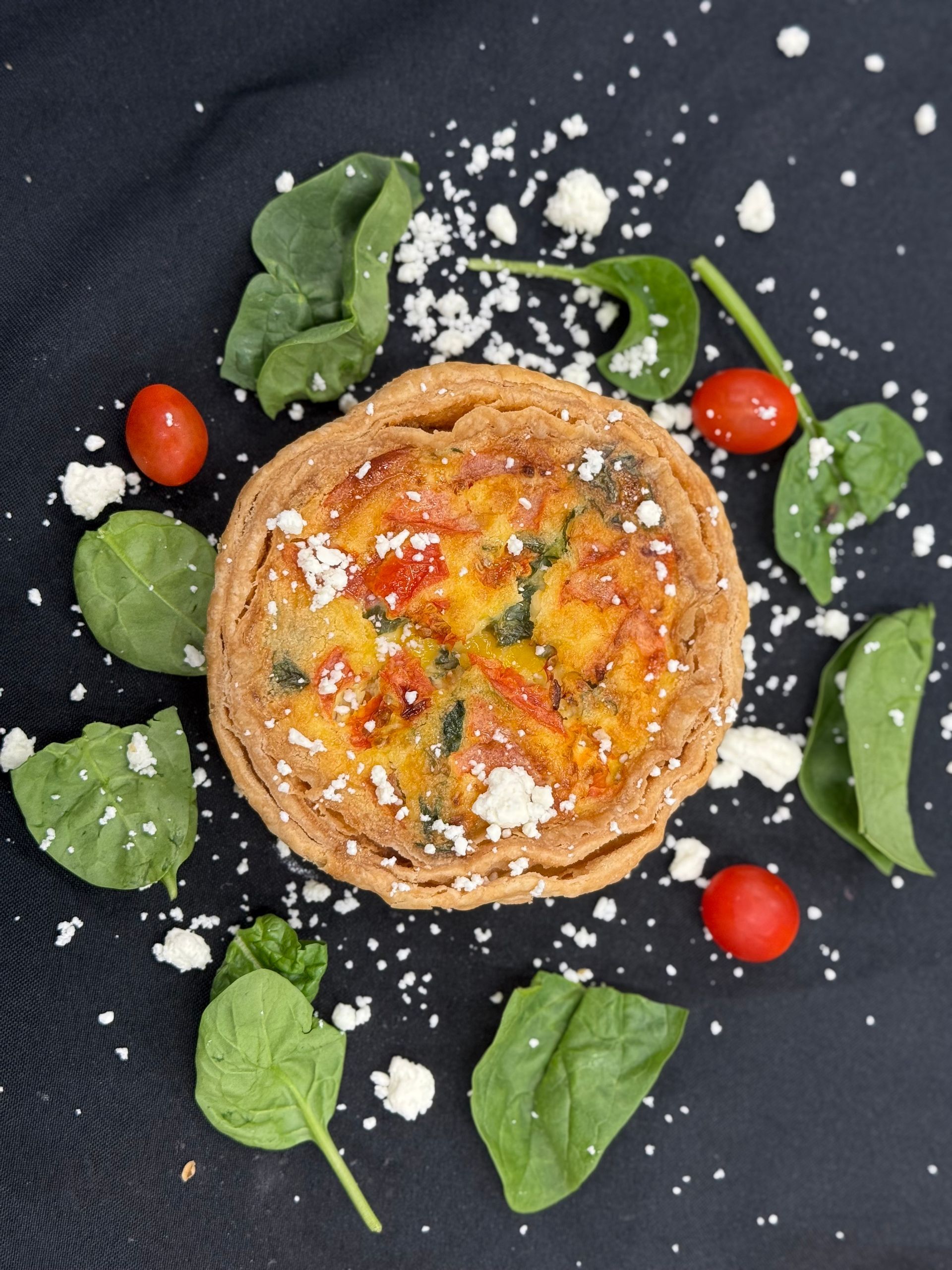 Quiche with spinach, tomatoes, and crumbled cheese on dark surface, surrounded by greens and tomatoes.
