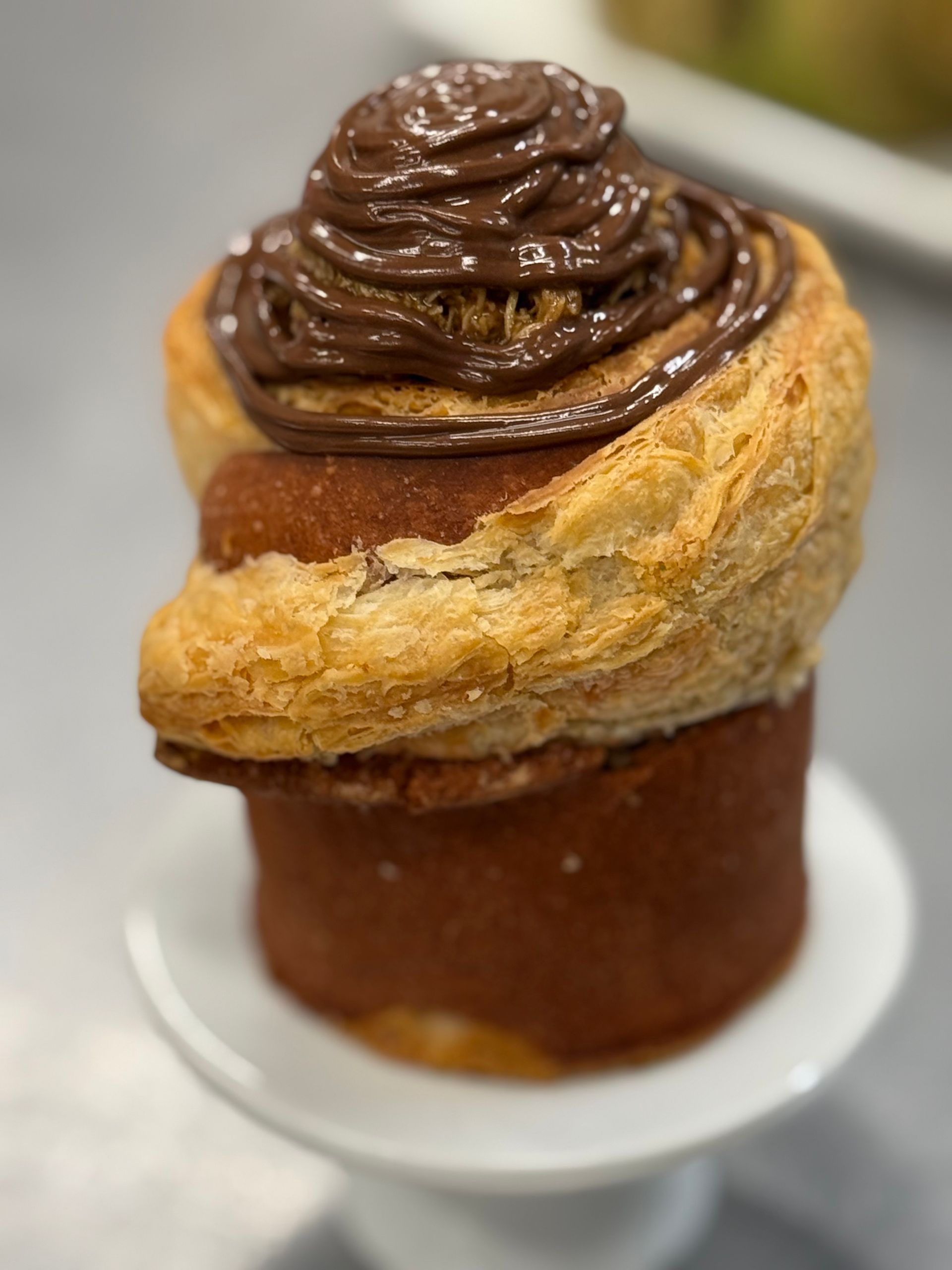 Cinnamon roll pastry with chocolate frosting, displayed on a small white pedestal.