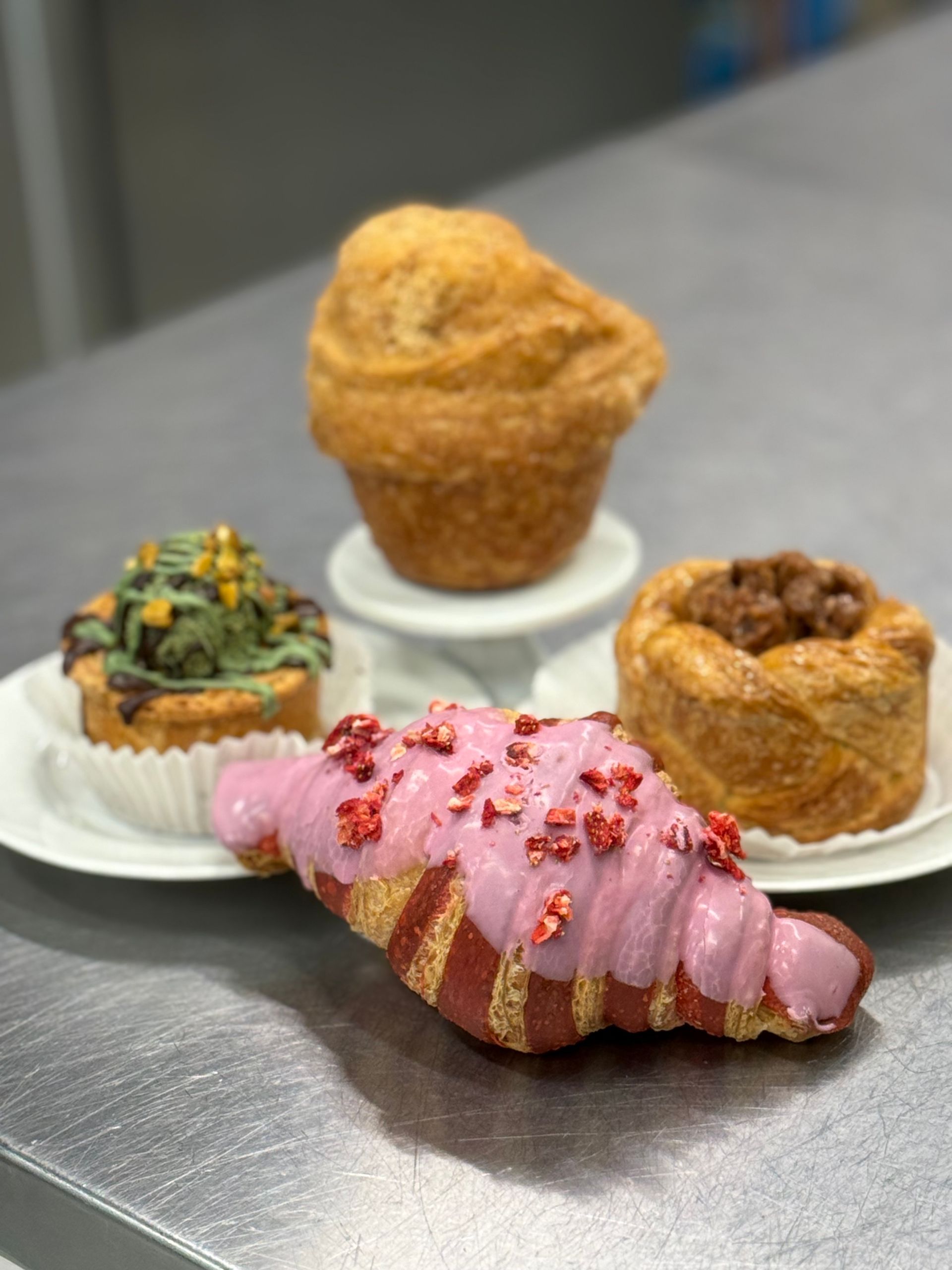Assortment of pastries on a metal surface: red-frosted croissant, green-topped tart, cylindrical pastry, and a muffin-shaped pastry.