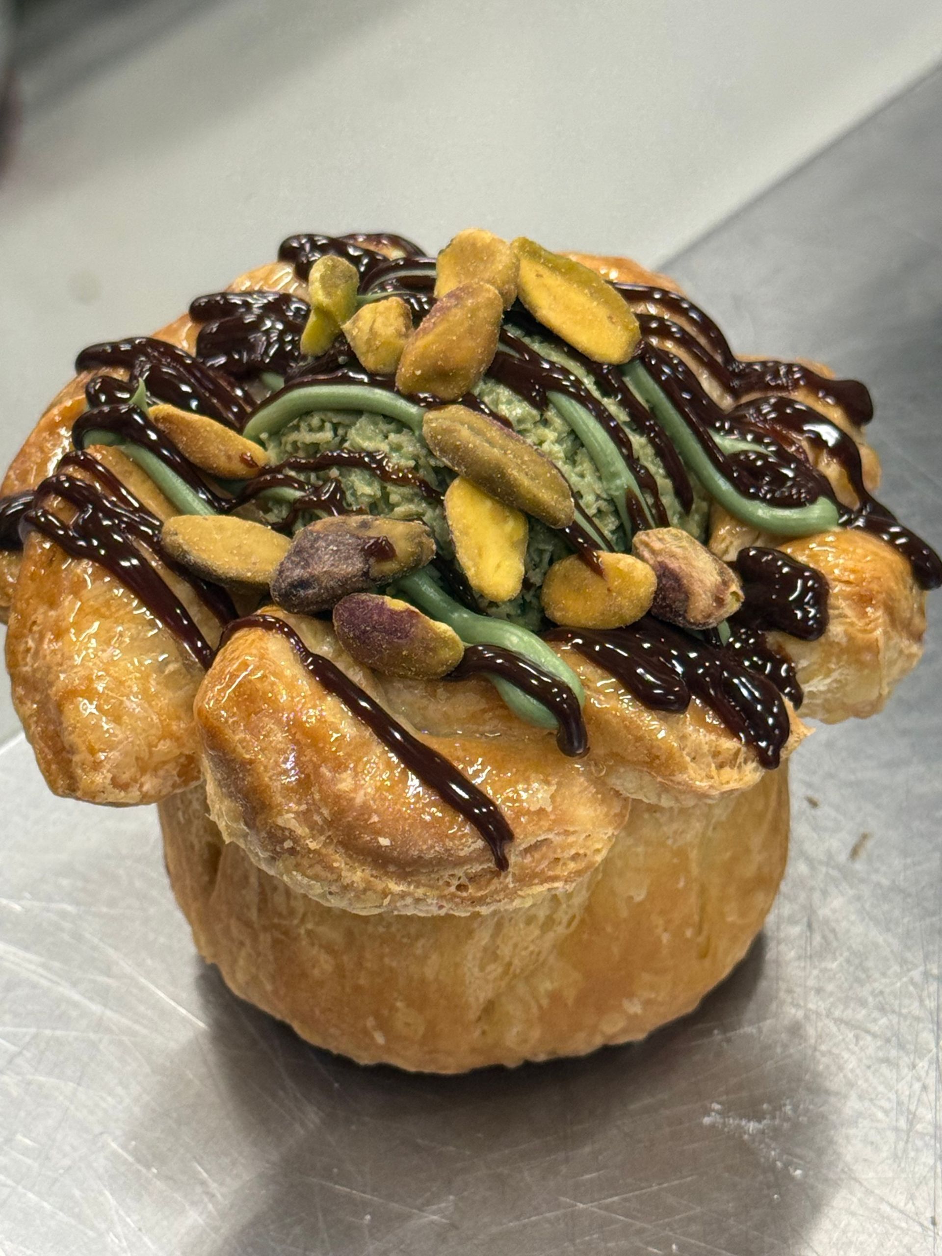 Pastry topped with green frosting, chocolate drizzle, and pistachios.