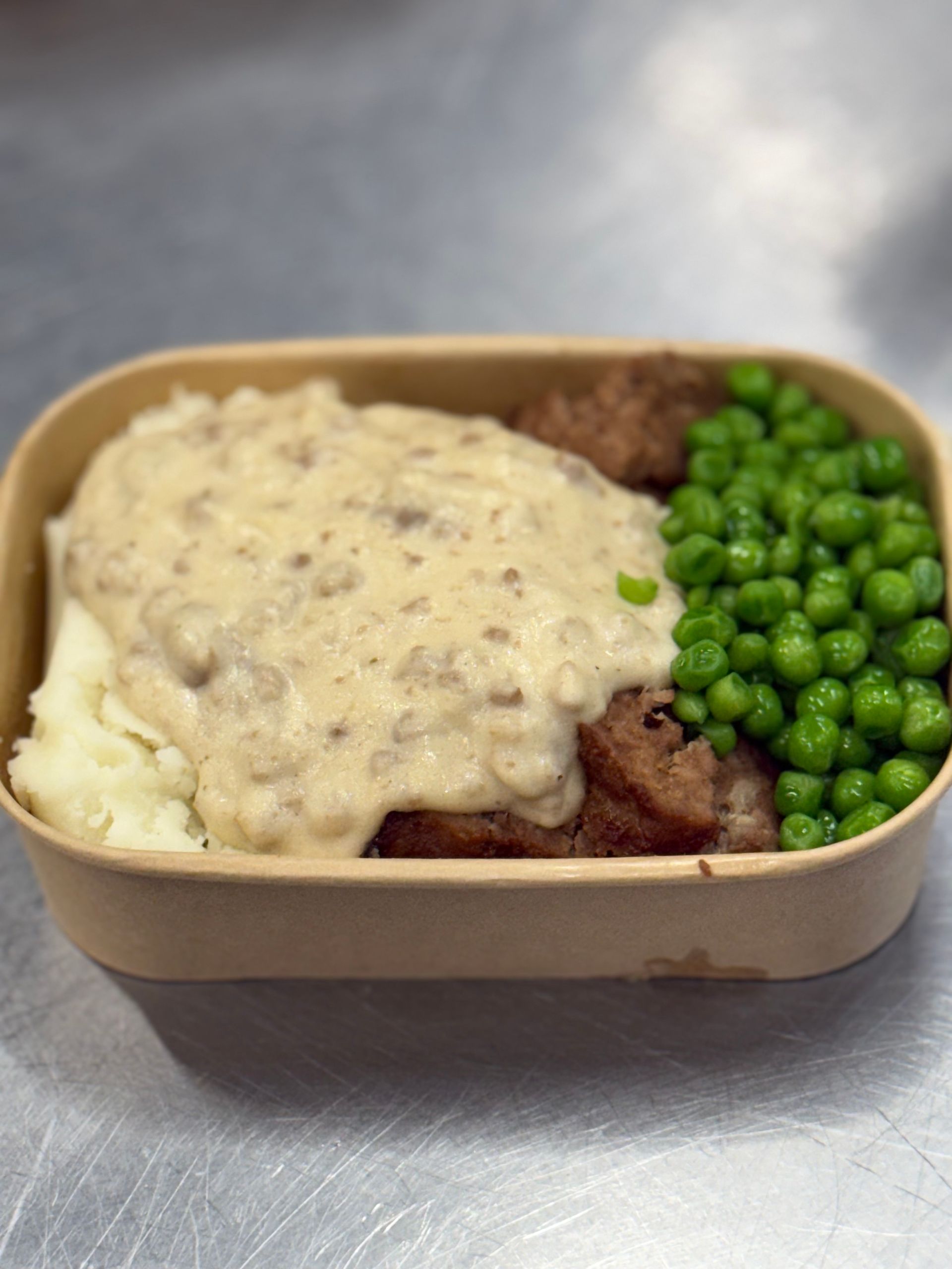 Meal of mashed potatoes, gravy, meatballs, and peas in a rectangular container.