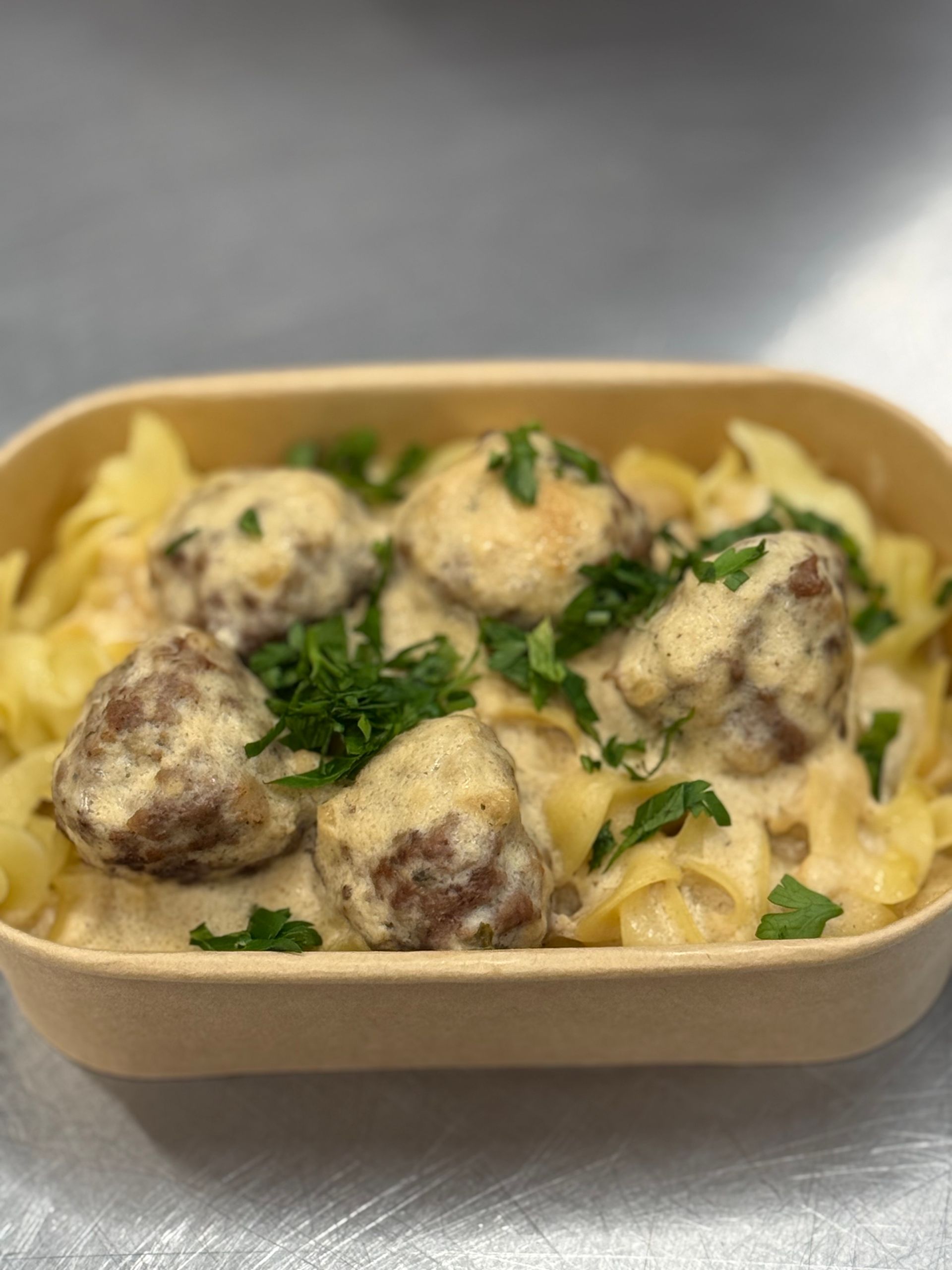 Meatballs in creamy sauce over noodles, garnished with parsley, served in a beige container.