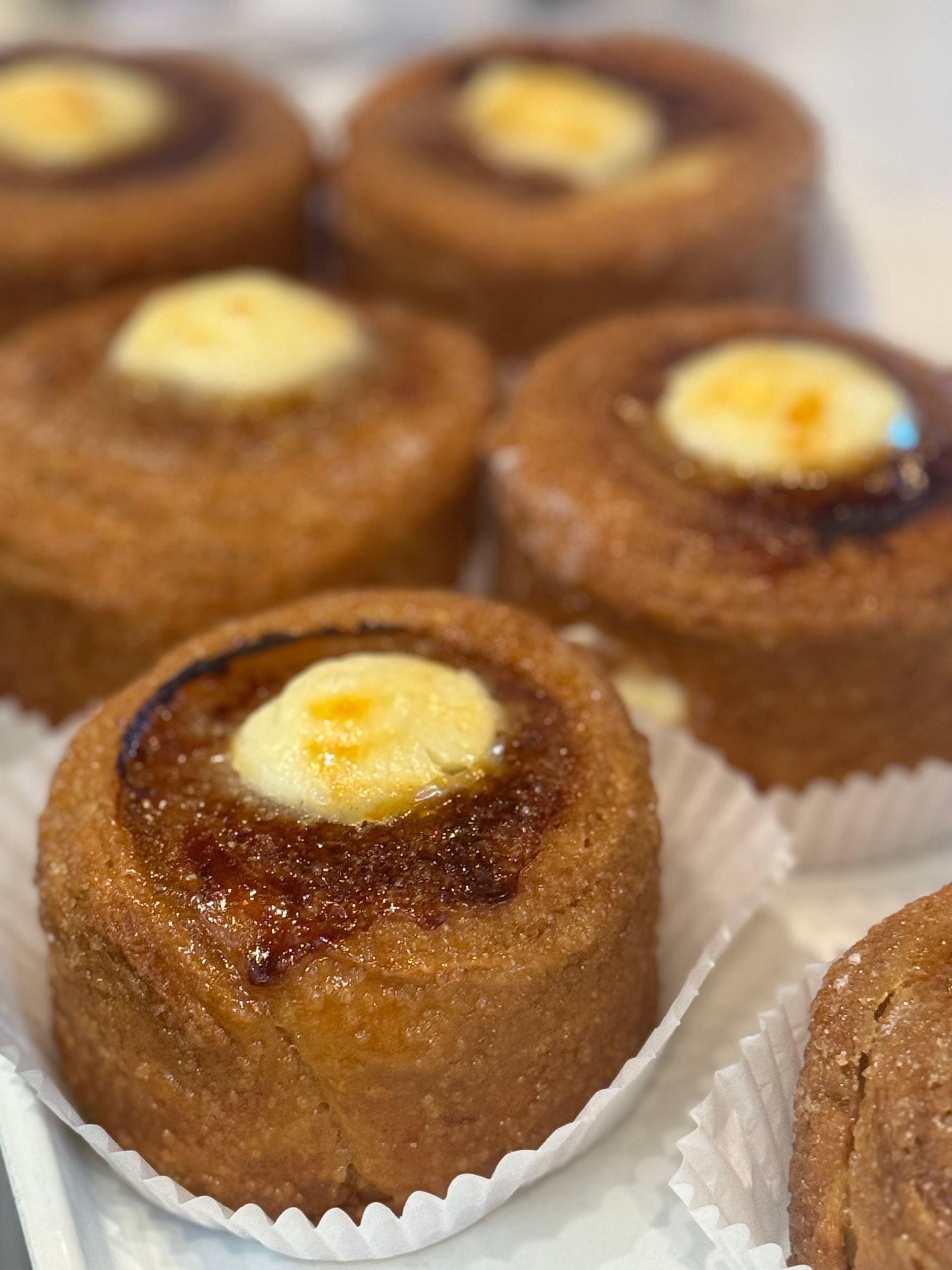 Close-up of several round, golden-brown pastries with a creamy topping and caramelized glaze, in paper cups.