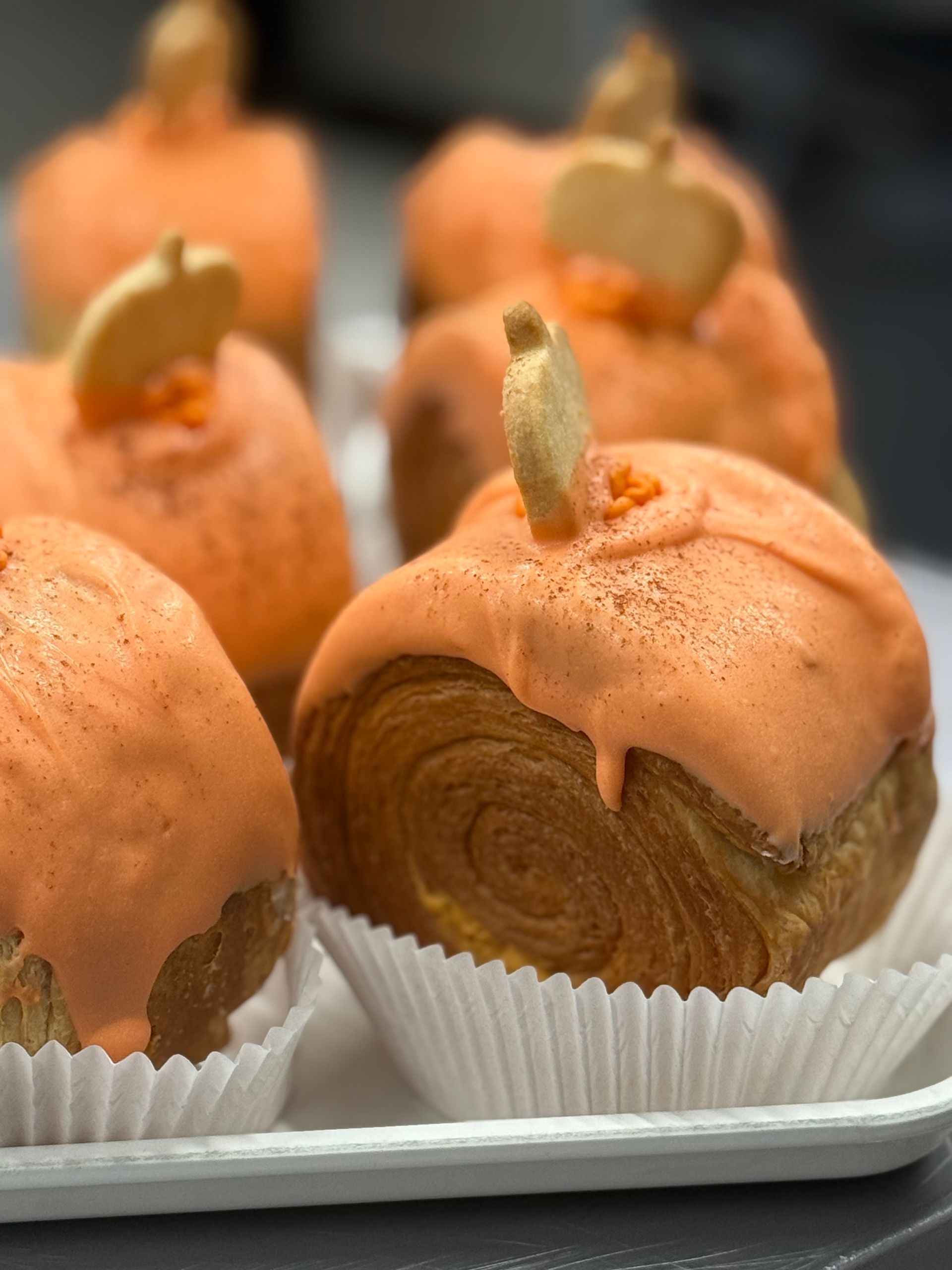 Pumpkin-shaped pastries in white paper cups, topped with orange icing and a cookie stem.