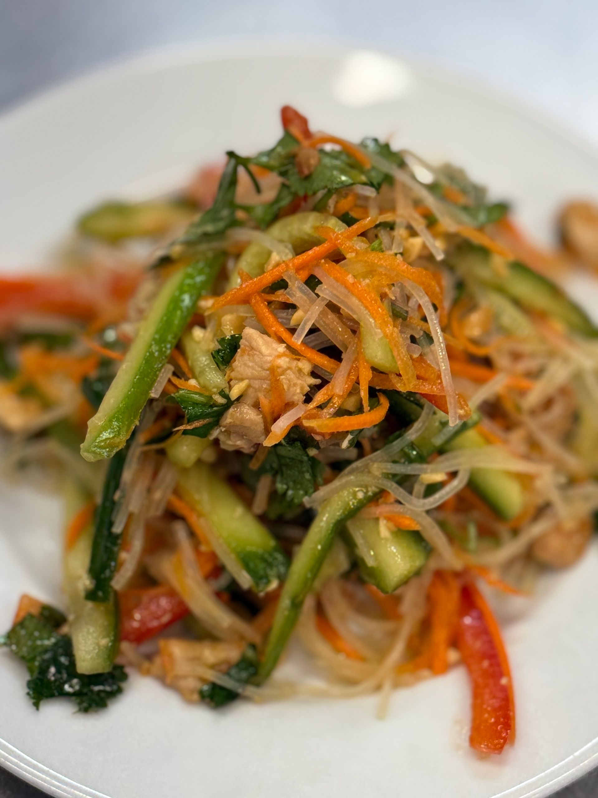 Colorful salad with noodles, cucumbers, carrots, herbs, and red peppers on a white plate.