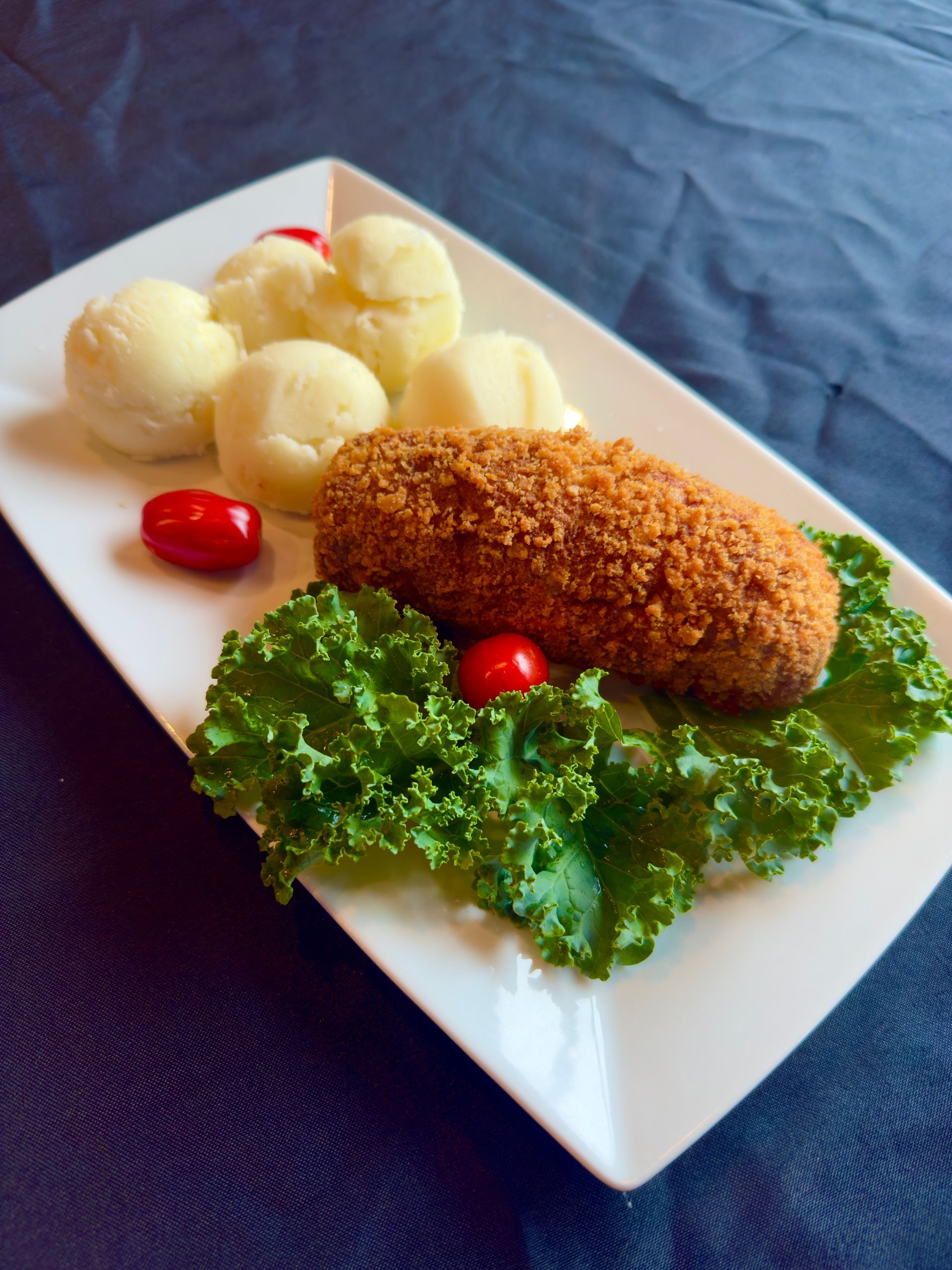 Plate with breaded food, potatoes, kale, and cherry tomatoes on a dark blue surface.