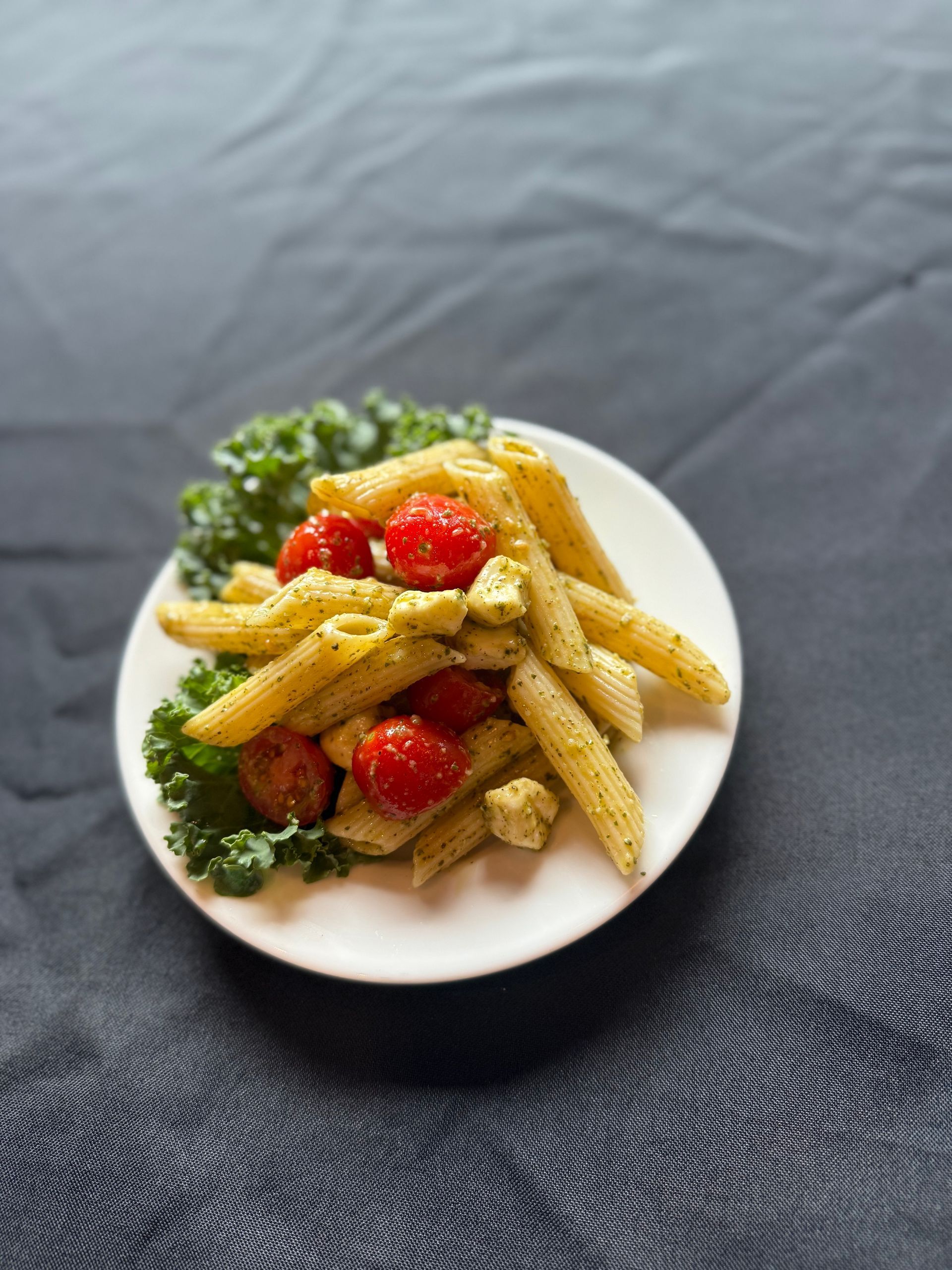 Pasta dish with cherry tomatoes and pesto, on a white plate with kale, on a dark fabric.