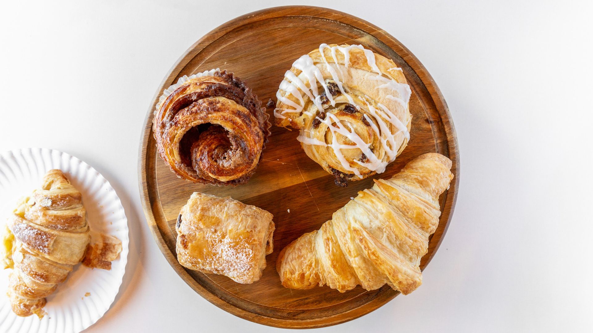 Pastries on a wooden tray and a paper plate: cinnamon roll, croissant, danish, and pain au chocolat.