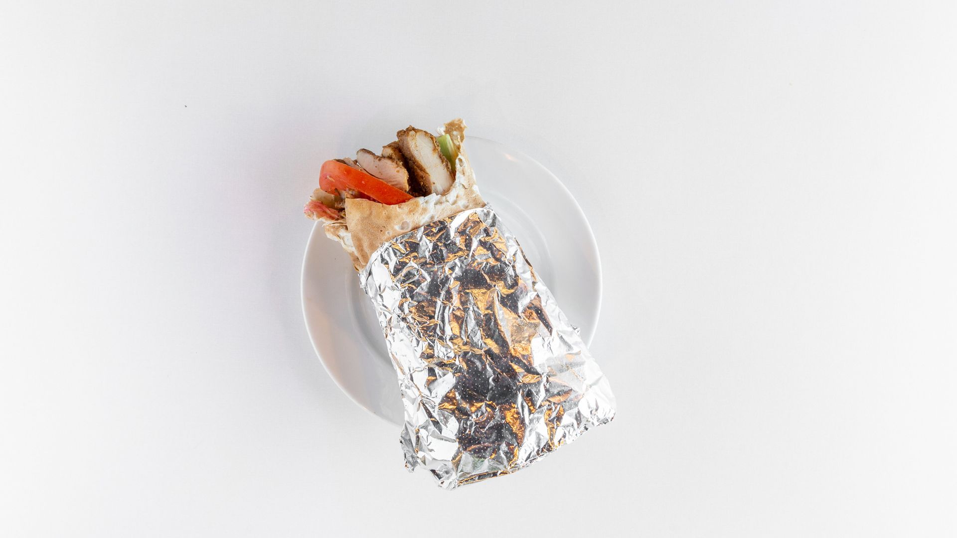 Shawarma wrap on a white plate, wrapped in foil. Visible: tomato, meat, and lettuce.
