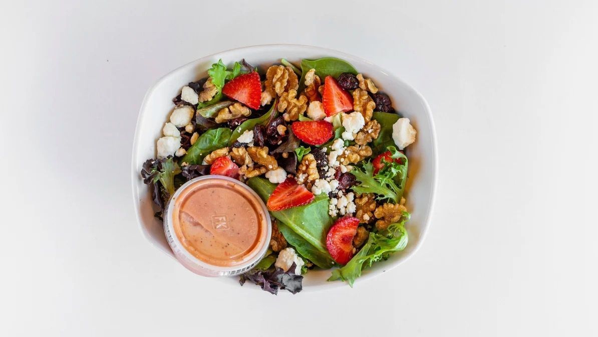 Salad with strawberries, walnuts, and feta cheese in a white bowl, dressing on the side.