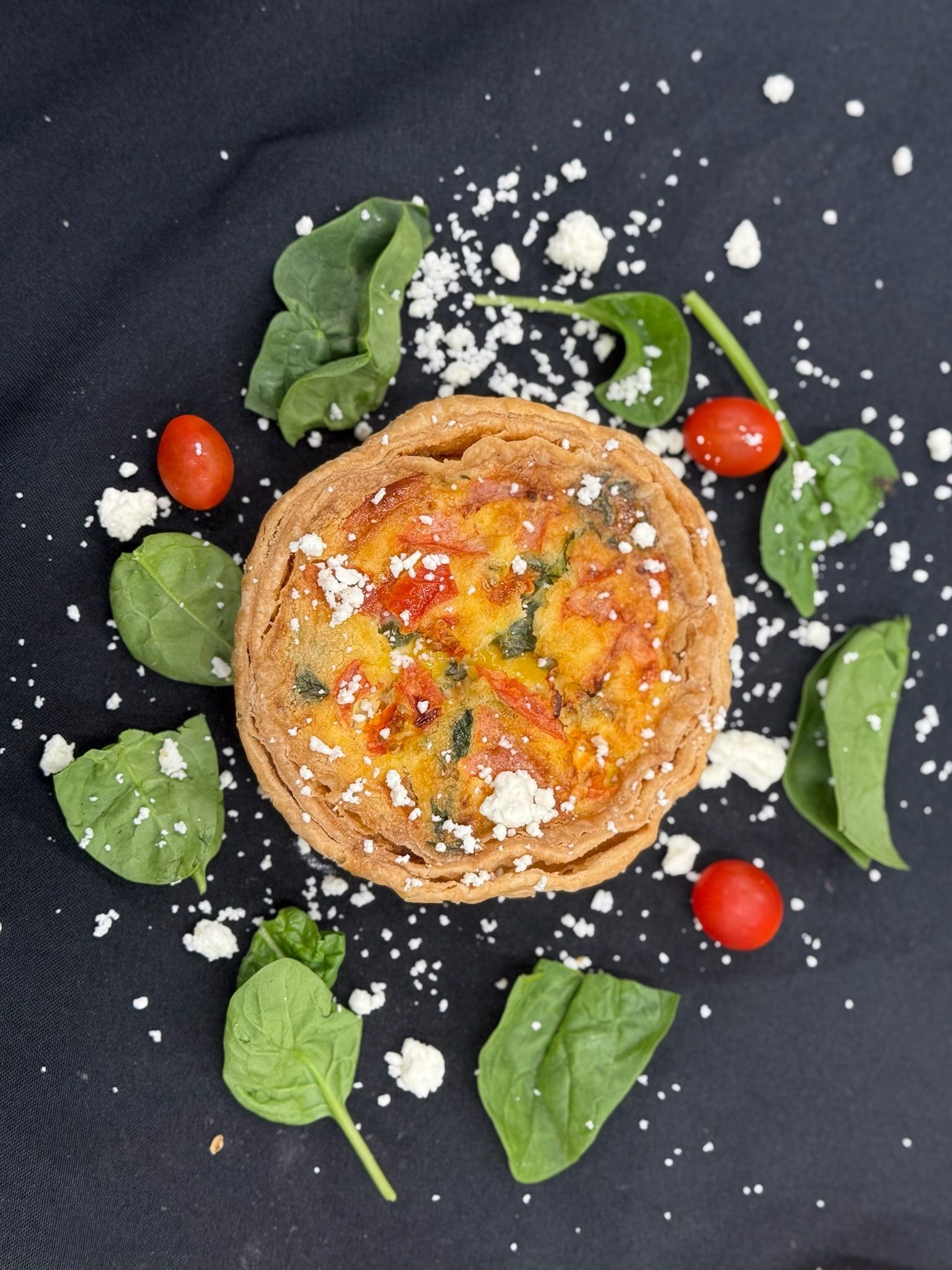 Quiche with tomatoes and spinach, surrounded by greens and crumbled cheese on a black surface.
