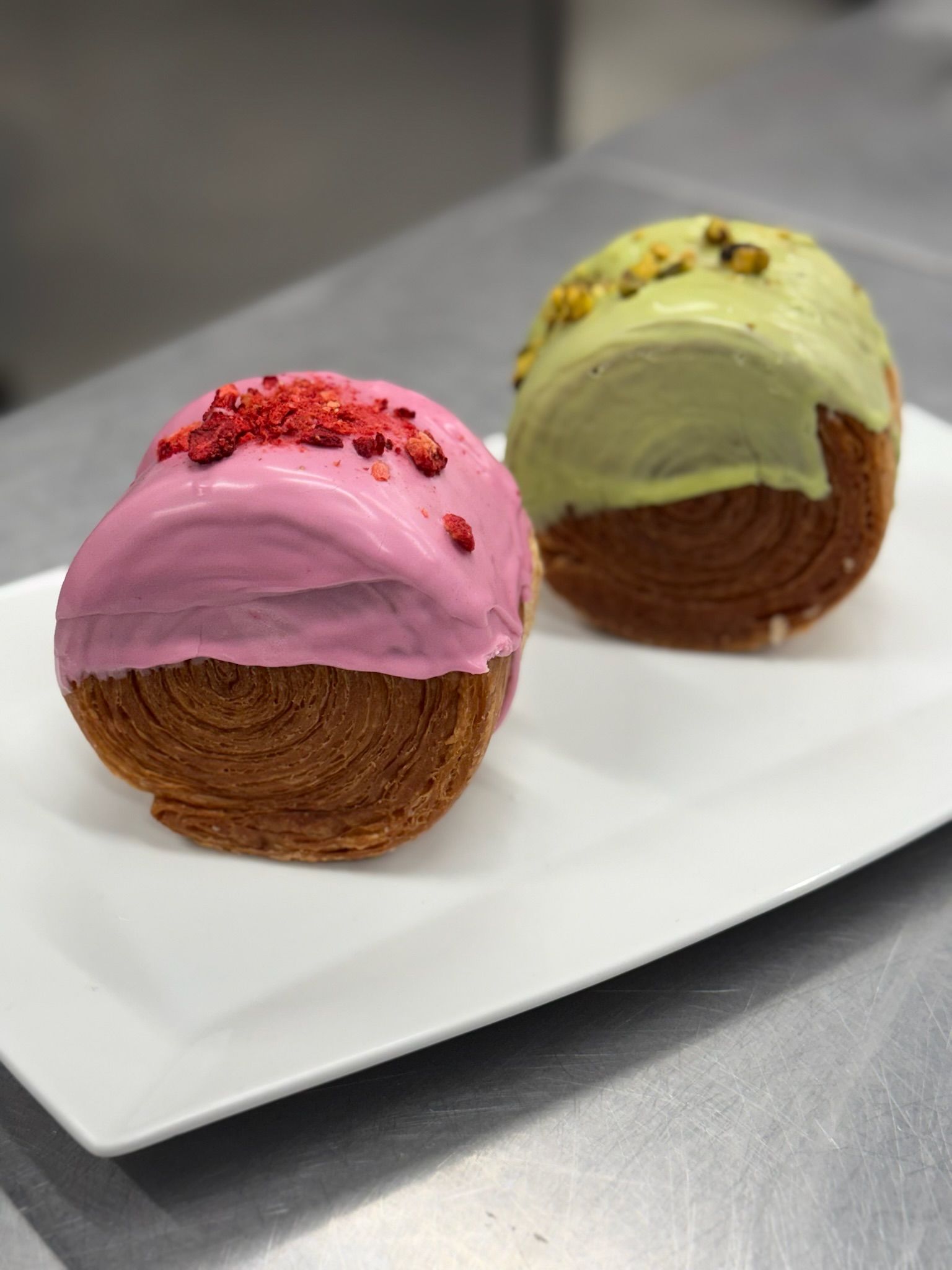 Two croissant-shaped pastries on a white plate, one with pink frosting, one with green frosting and pistachios.