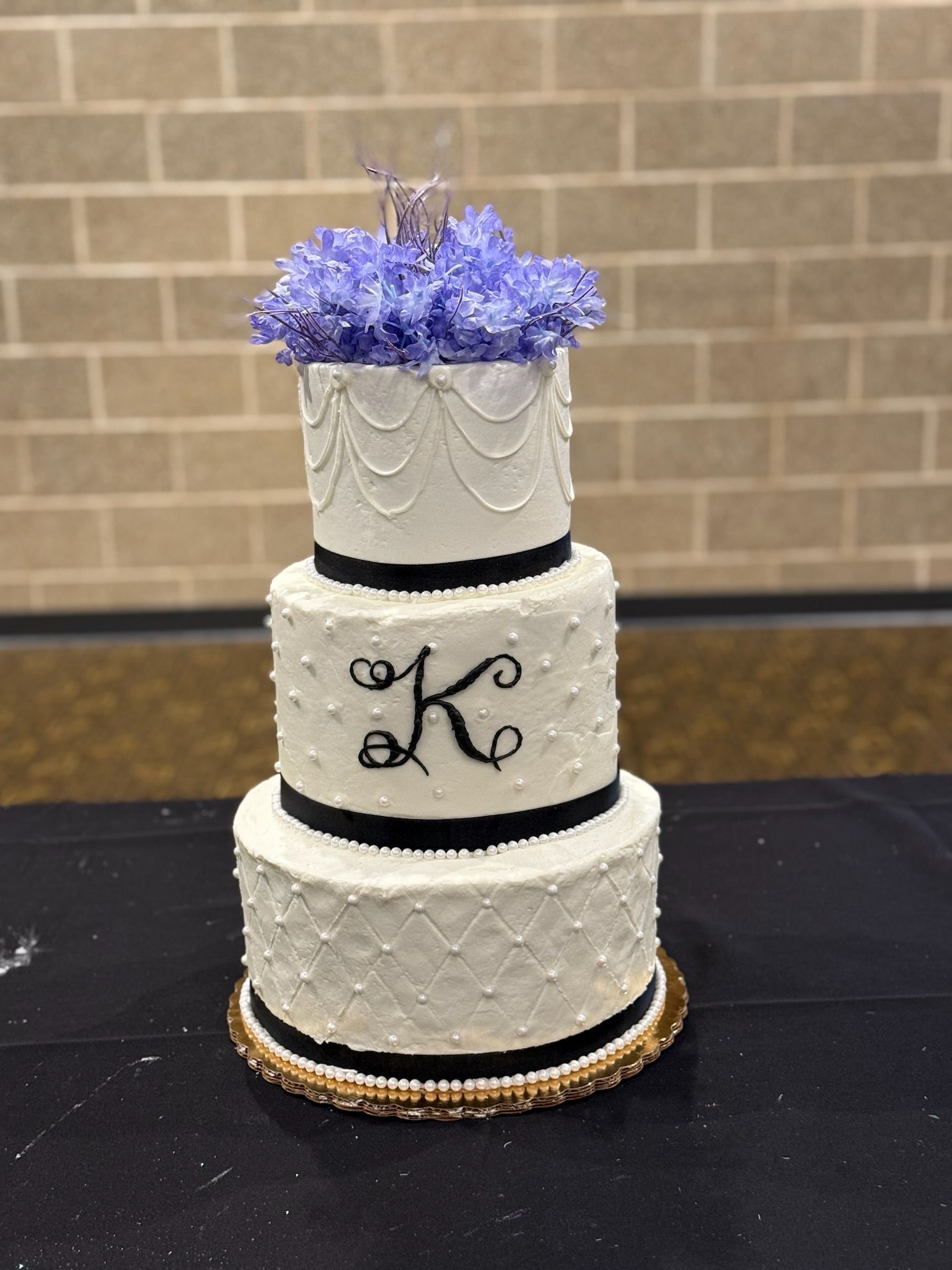 Three-tier white cake with black accents and a purple floral topper, on a black tablecloth, against a brick wall.