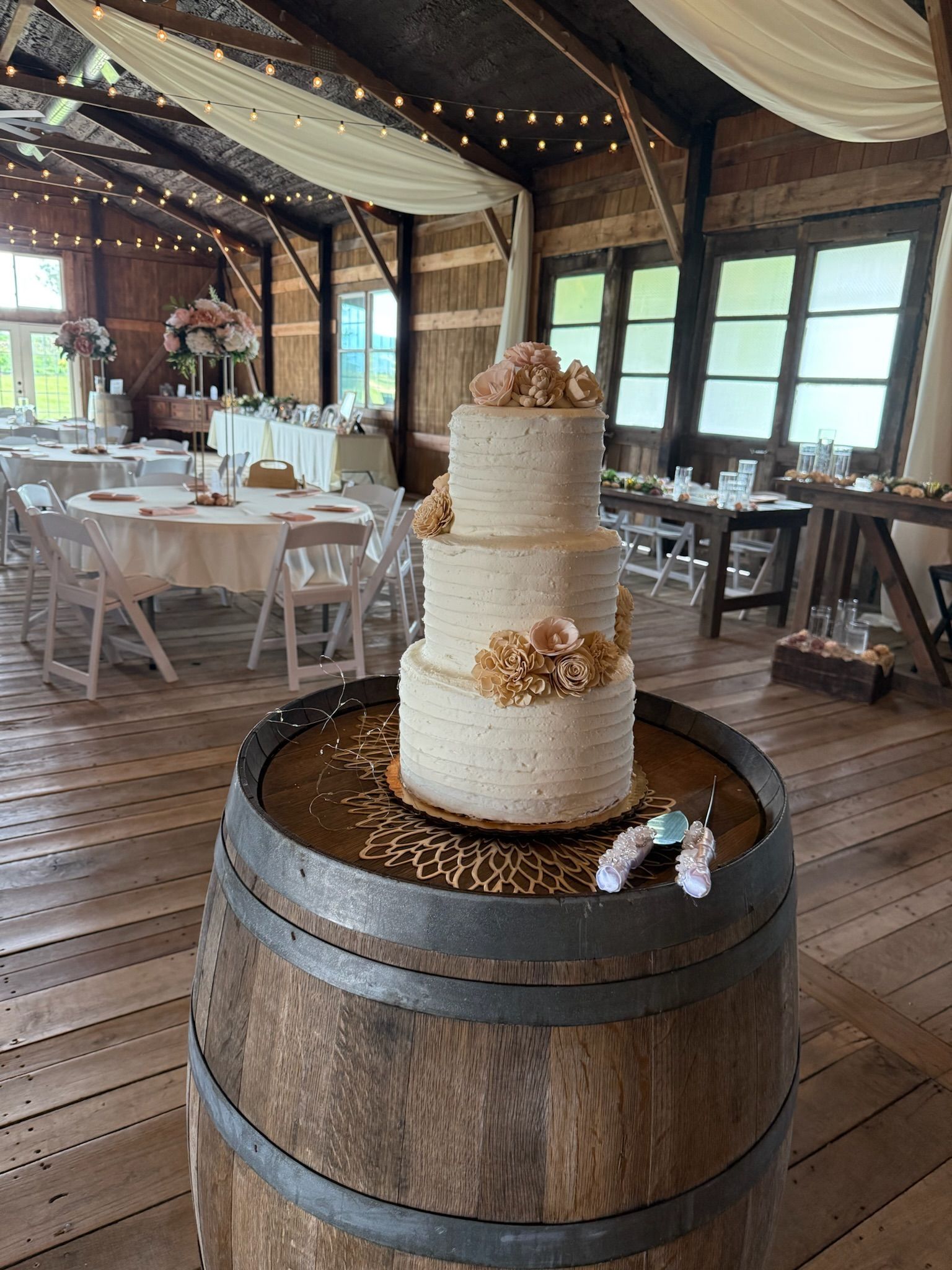 Three-tiered wedding cake with floral accents sits on a barrel in a rustic barn setting with tables.